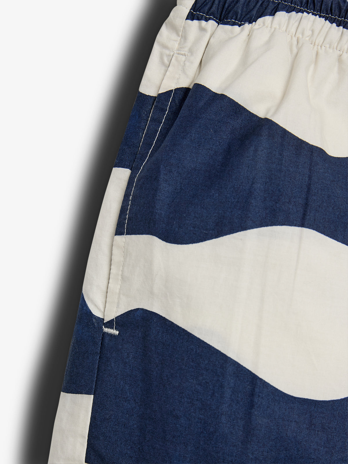 Die Nahaufnahme zeigt einen Abschnitt eines Kleidungsstücks mit blau-weißen Wellenmustern. Der Stoff ist elastisch an der oberen Naht zusammengezogen und hat eine Seitentasche.
