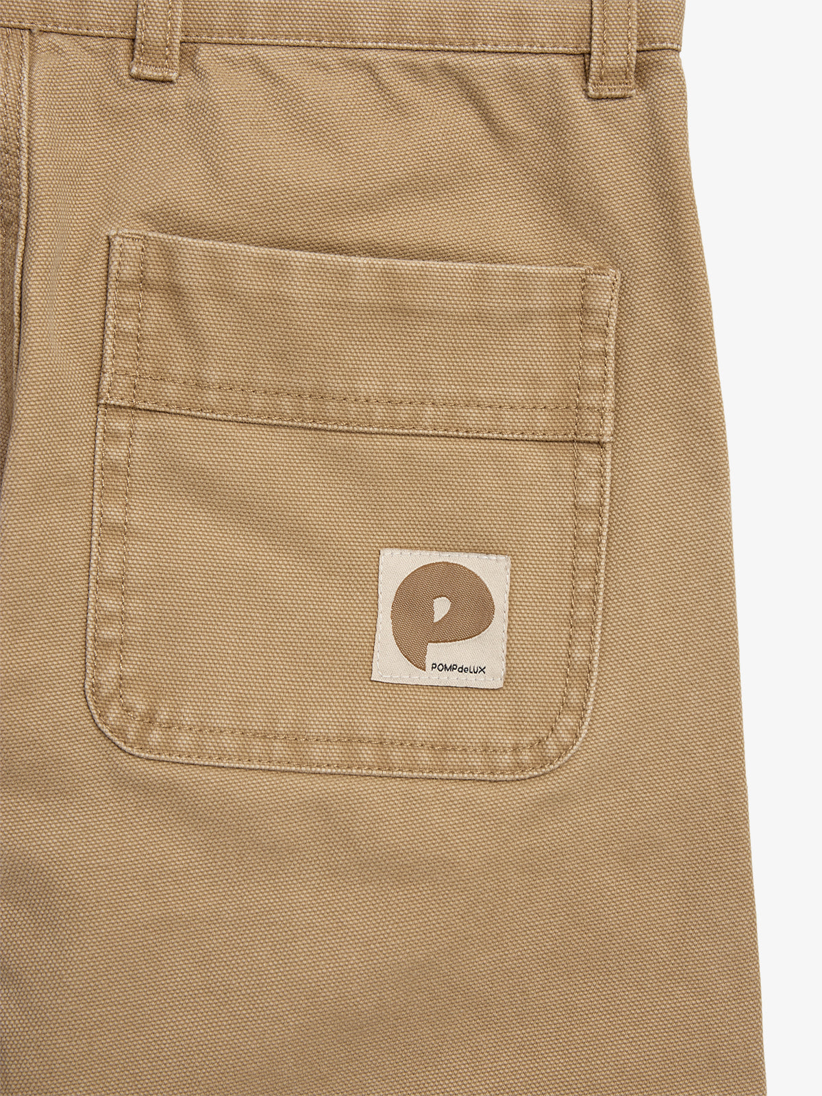 Eine beige Tasche ist auf einem Kleidungsstück zu sehen, mit einem Label, das ein braunes "P" und den Text "POMPdeLUX" zeigt. Umgeben von Stoff und Gürtelschlaufen.