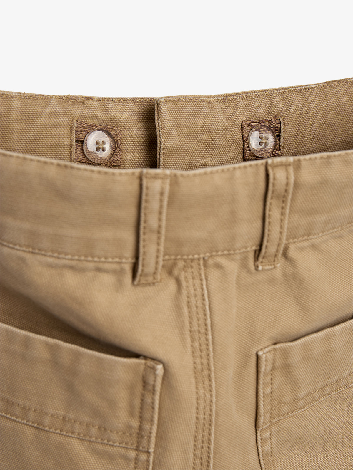 Eine beige Hose zeigt den oberen Bund mit zwei weißen Knöpfen befestigt. Zwei große Vordertaschen sind sichtbar. Der Hintergrund ist neutral gehalten.