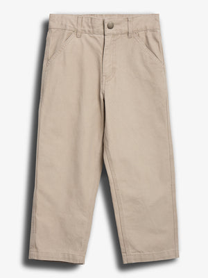 Beige Kinderhose liegt flach mit zwei seitlichen Taschen, Reißverschluss und Knopf. Kontext ist ein neutraler Hintergrund, der die Hose in den Fokus rückt. Keine Textbeschriftung vorhanden.