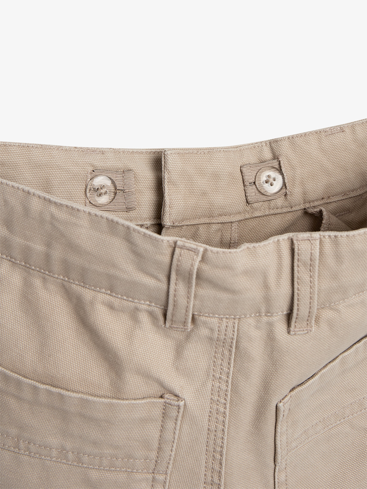 Beige Hose mit Knöpfen und Gürtelschlaufen sichtbar, liegt flach. Der Kontext zeigt den oberen Teil der Hose vor einem einfachen, hellen Hintergrund.