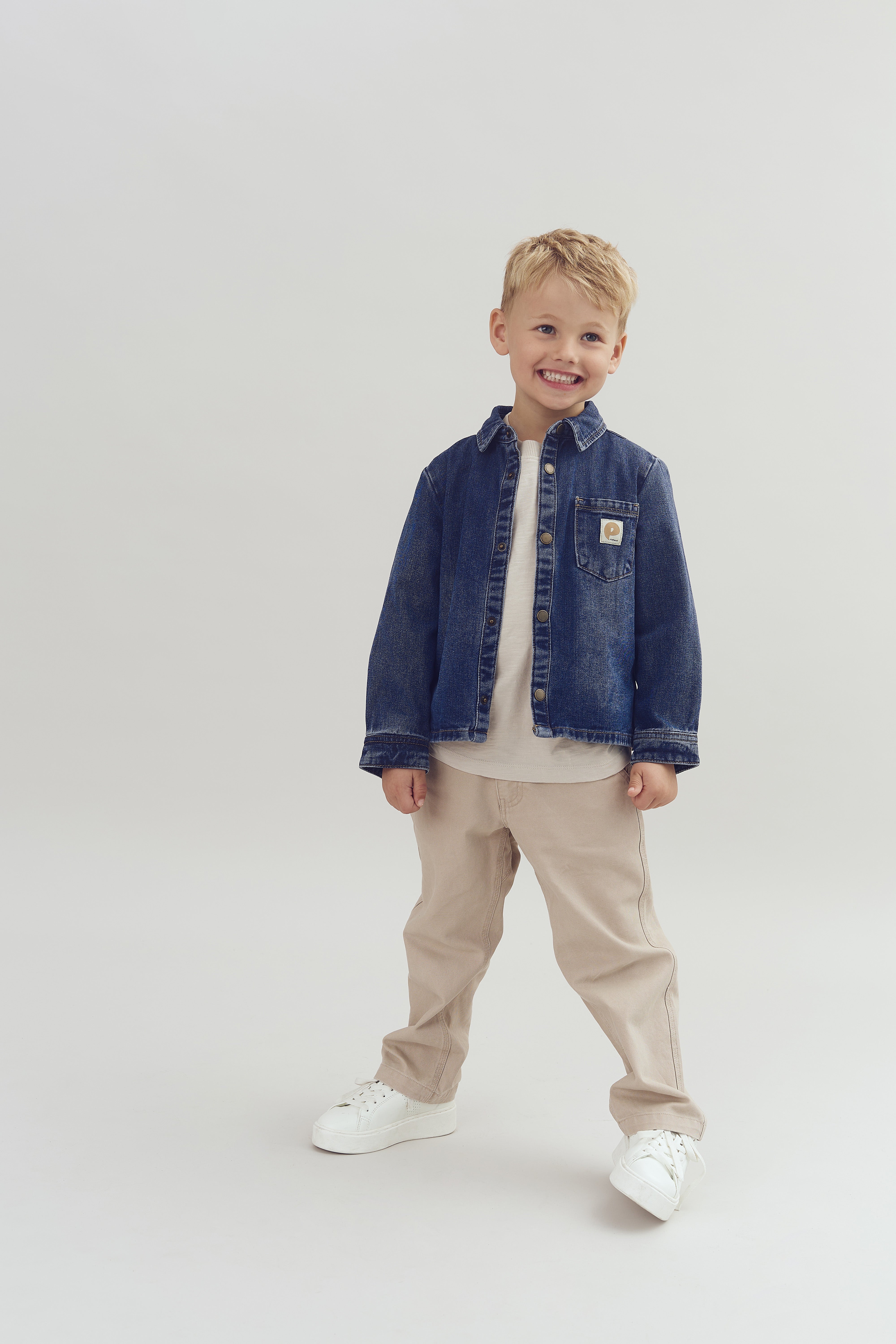 Ein Kind lächelt in die Kamera, trägt eine blaue Jeansjacke, beige Hose und weiße Turnschuhe. Es steht auf einem neutralen Hintergrund.