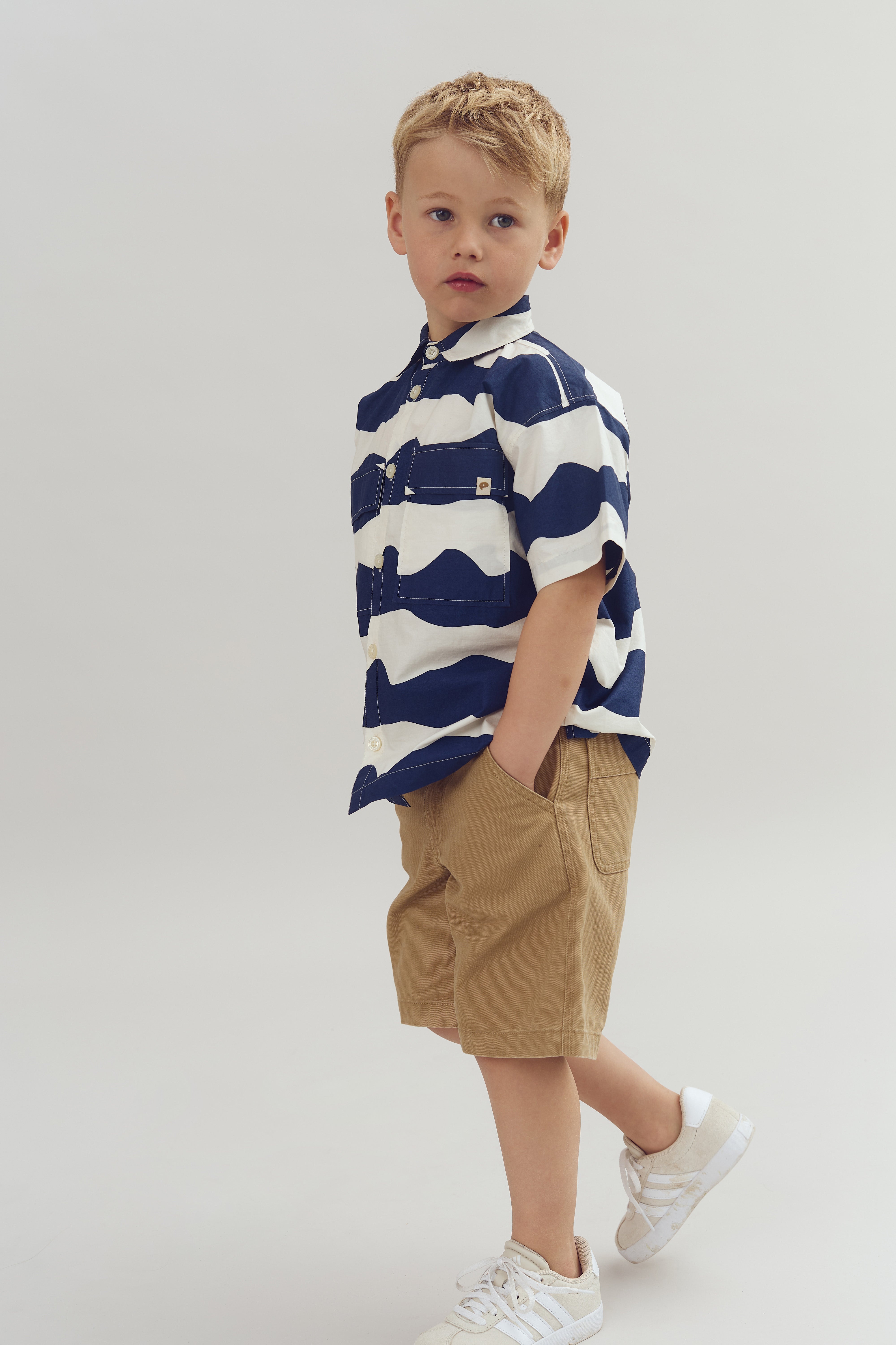 Ein kleiner Junge steht mit den Händen in den Taschen. Er trägt ein gestreiftes Hemd und beige Shorts. Der Hintergrund ist neutral und hell.