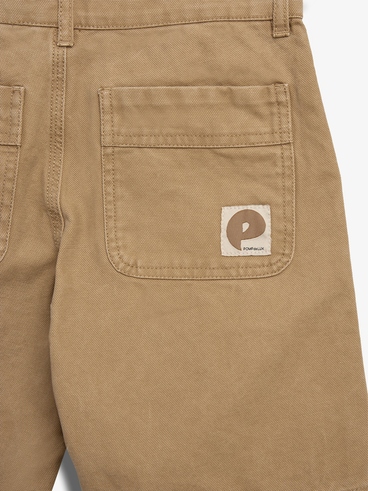 Beige Hose zeigt eine Gesäßtasche mit einem rechteckigen Etikett, das ein stilisiertes "P"-Logo und die Aufschrift "POMPdeLUX" enthält. Die Umgebung ist schlicht und einfarbig.