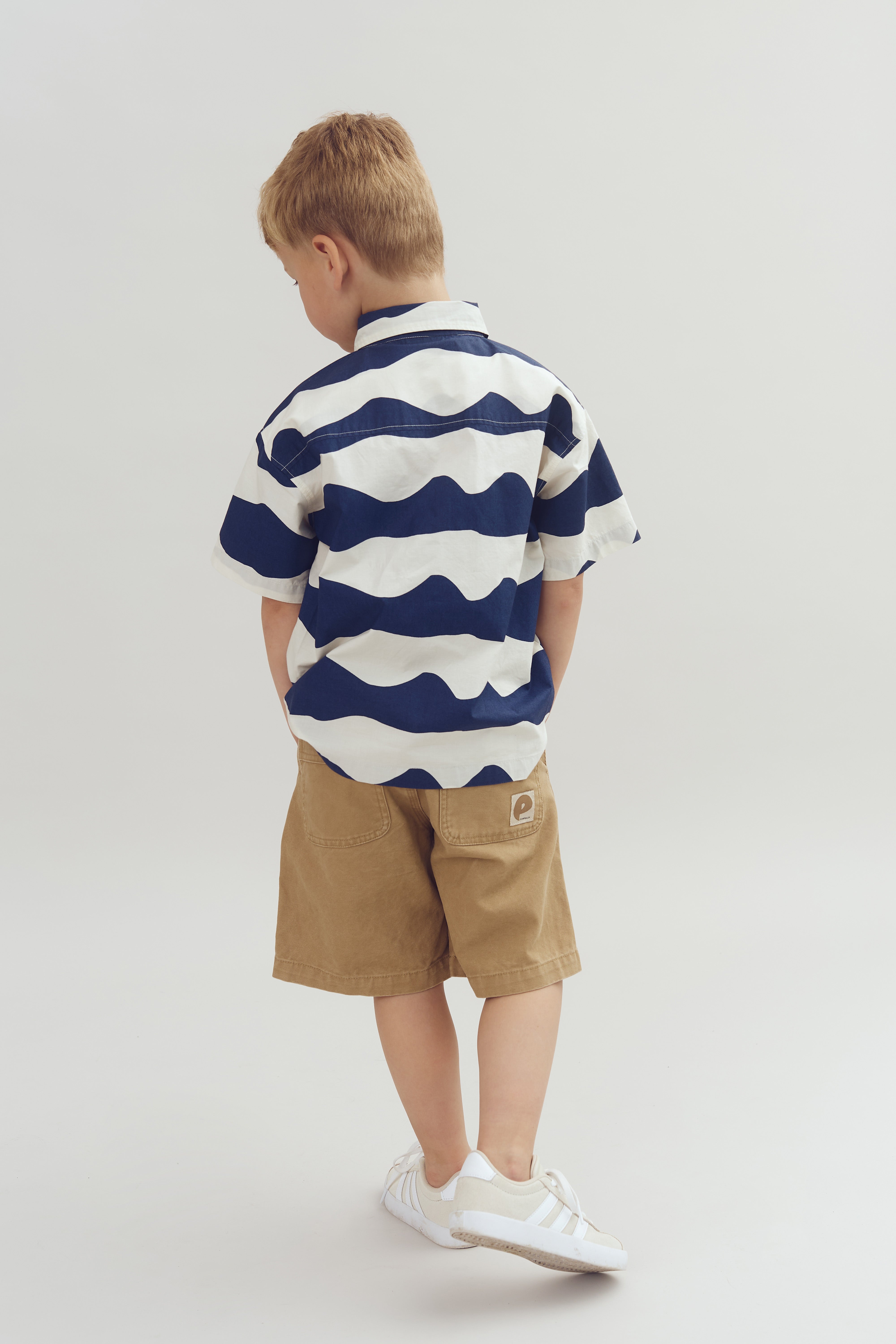 Ein Junge, gekleidet in ein blau-weiß gestreiftes Hemd und braune Shorts, steht mit dem Rücken zur Kamera in einem neutralen, grauen Raum.