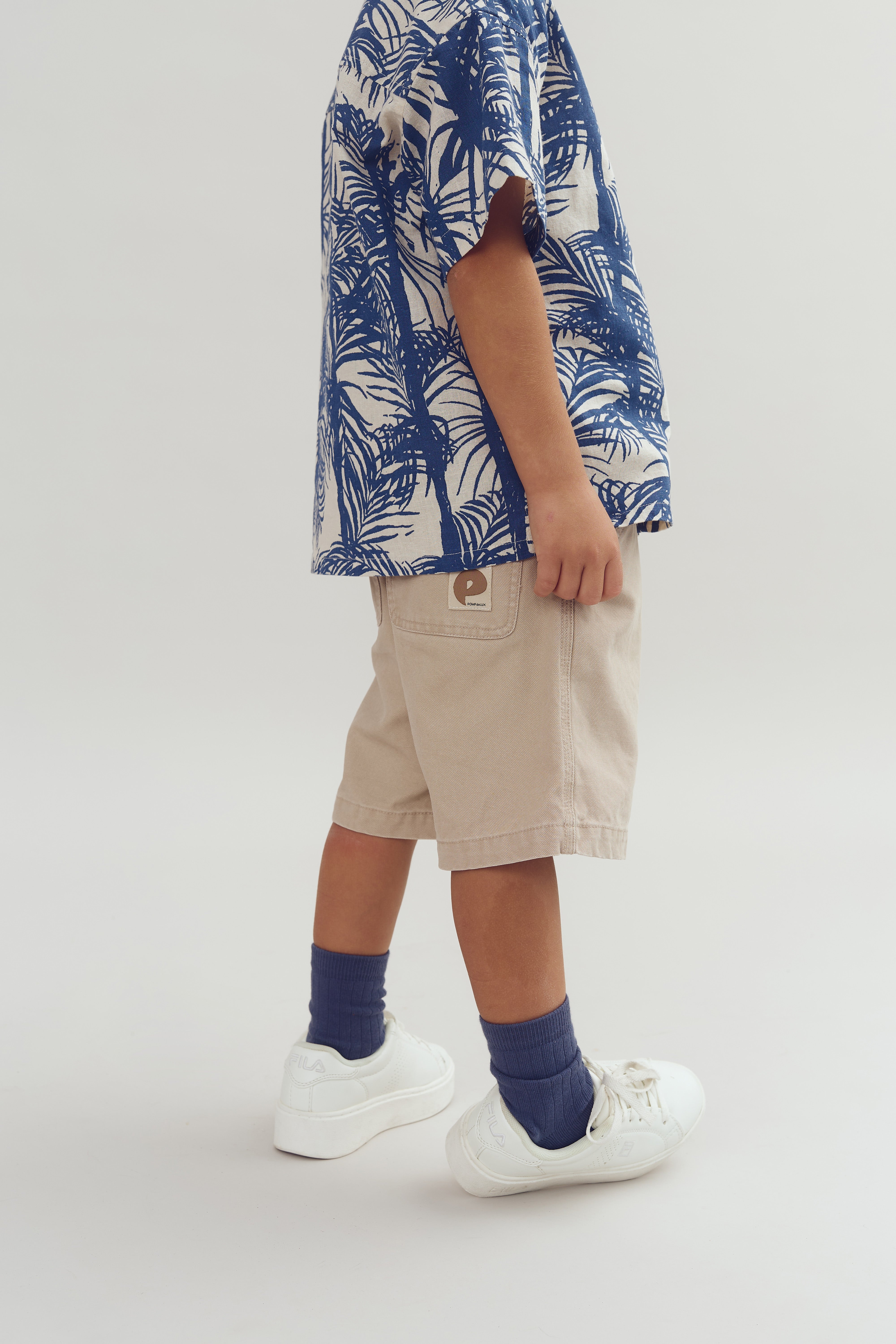 Ein Kind trägt ein blau-weiß gemustertes Hemd und beige Shorts. Dazu blaue Socken und weiße Turnschuhe auf einem neutralen Hintergrund.