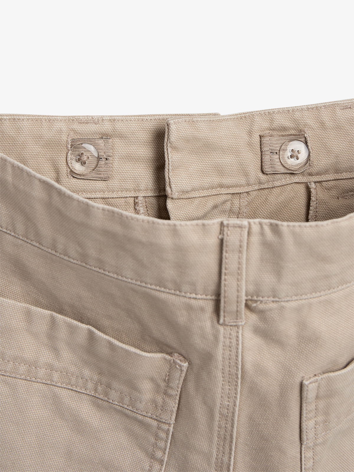 Eine beige Hose mit zwei sichtbaren Knöpfen und Gürtelschlaufen, in Nahaufnahme fotografiert, zeigt den oberen Bund und Teile der Gesäßtaschen. Der Hintergrund ist neutral.