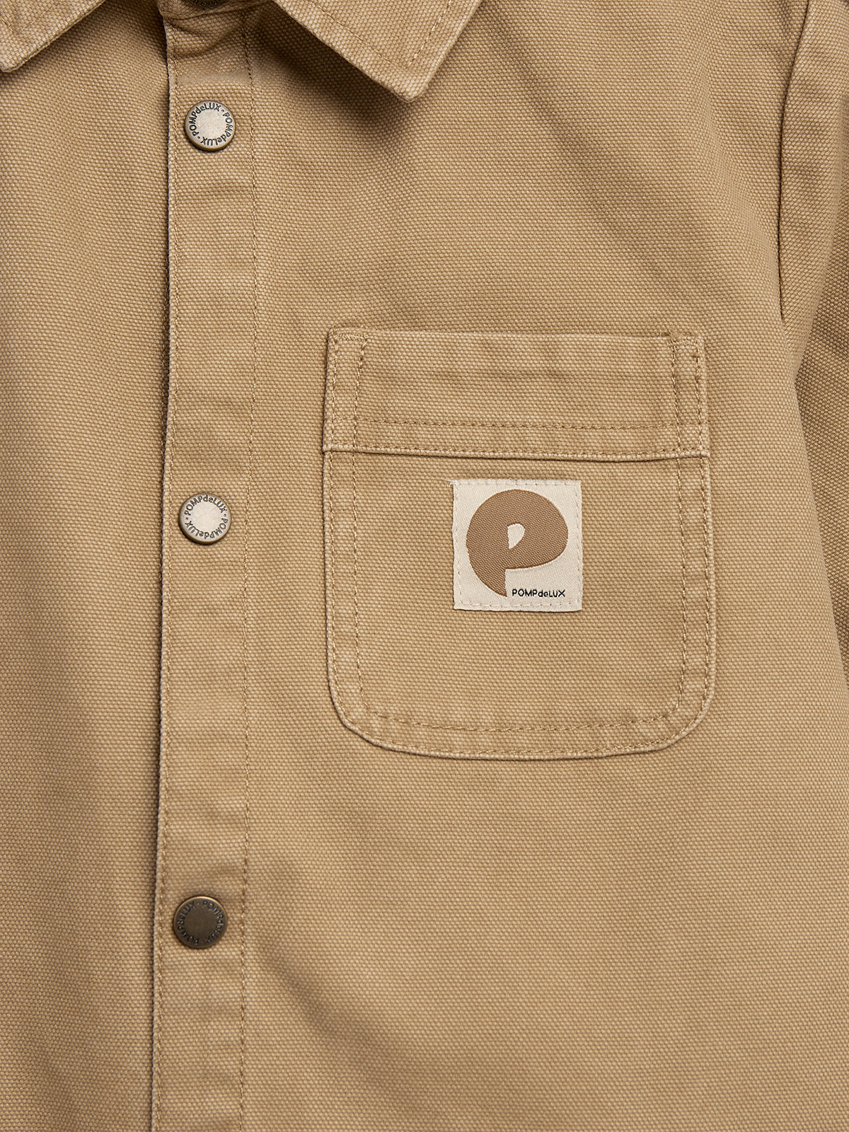 Ein beiges Hemd mit silbernen Druckknöpfen und einer Brusttasche. Auf der Tasche befindet sich ein Etikett mit einem Logo und dem Text "POM d'API POMME dUX".