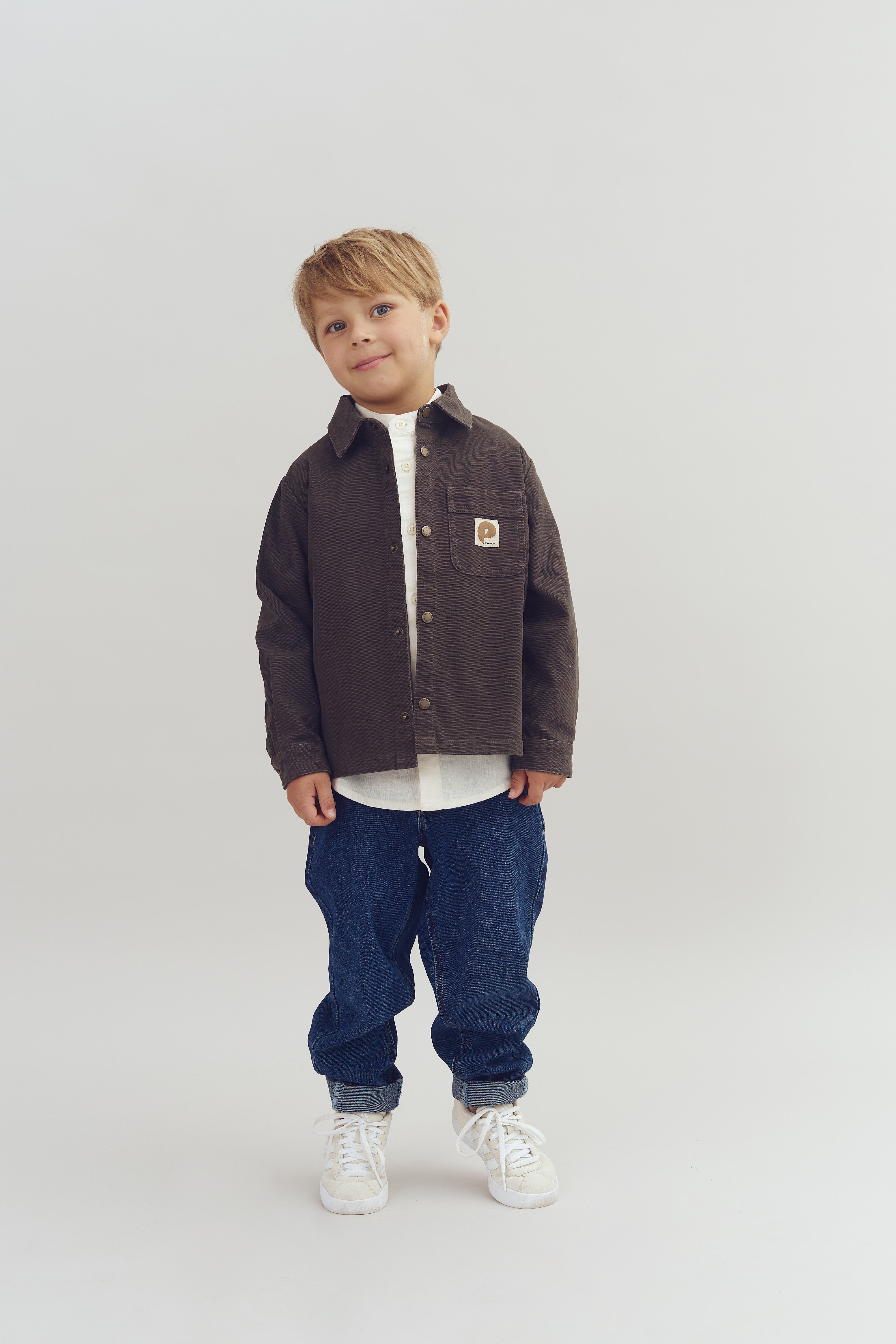 Ein Junge trägt ein braunes Hemd mit Logo, steht ruhig in der Mitte eines hellen, neutralen Hintergrunds. Die Ärmel sind locker, blaue Jeans und weiße Turnschuhe vervollständigen das Outfit.