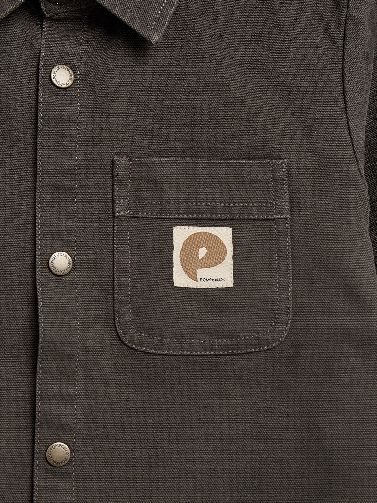 A close-up of a dark button-up shirt with metal buttons and a chest pocket. The pocket features a beige tag with a brown circular logo and the text "POMPdeLUX."
