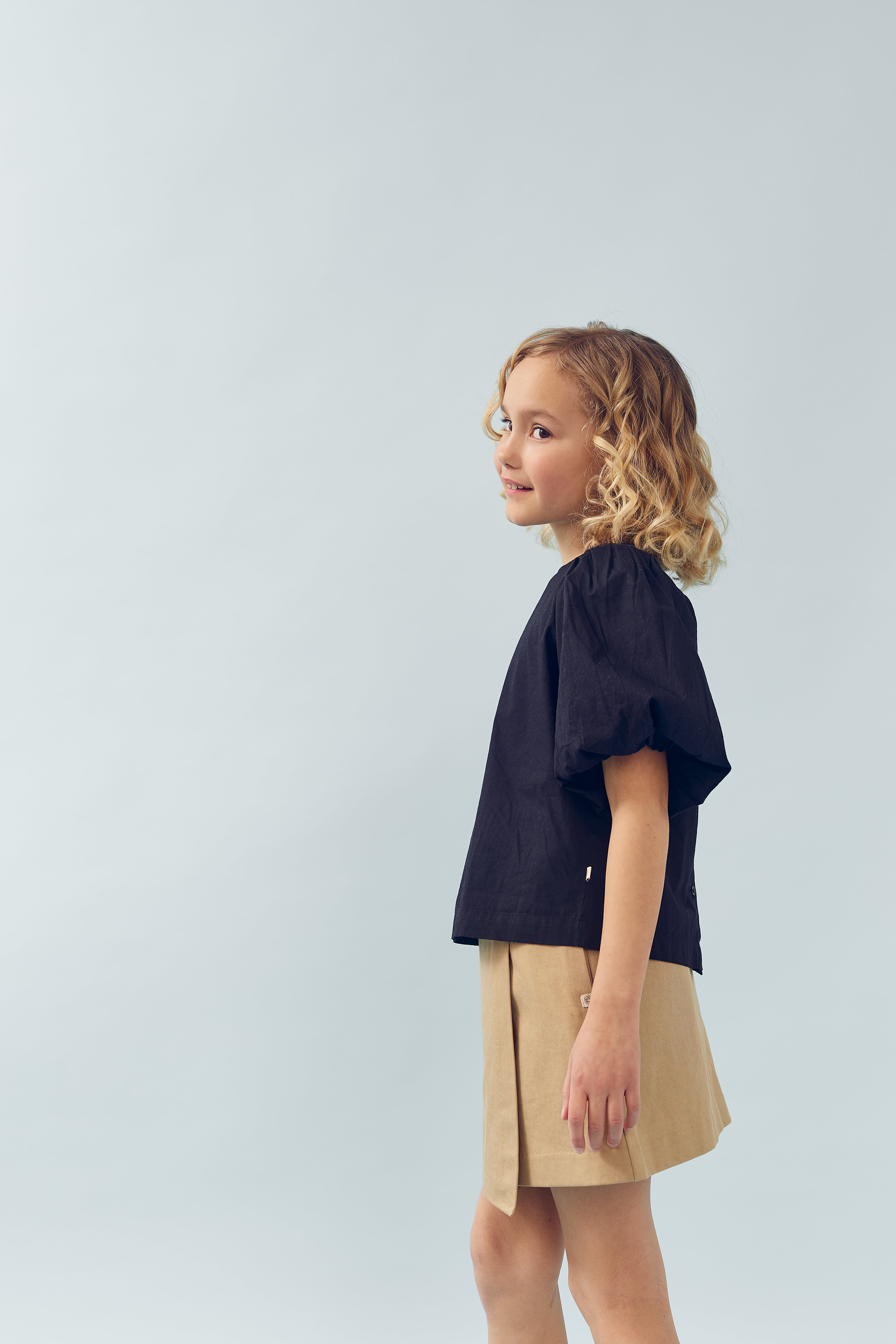 Ein Kind mit lockigem Haar steht seitlich, trägt ein schwarzes Shirt und einen beigefarbenen Rock, vor einem hellgrauen, neutralen Hintergrund.