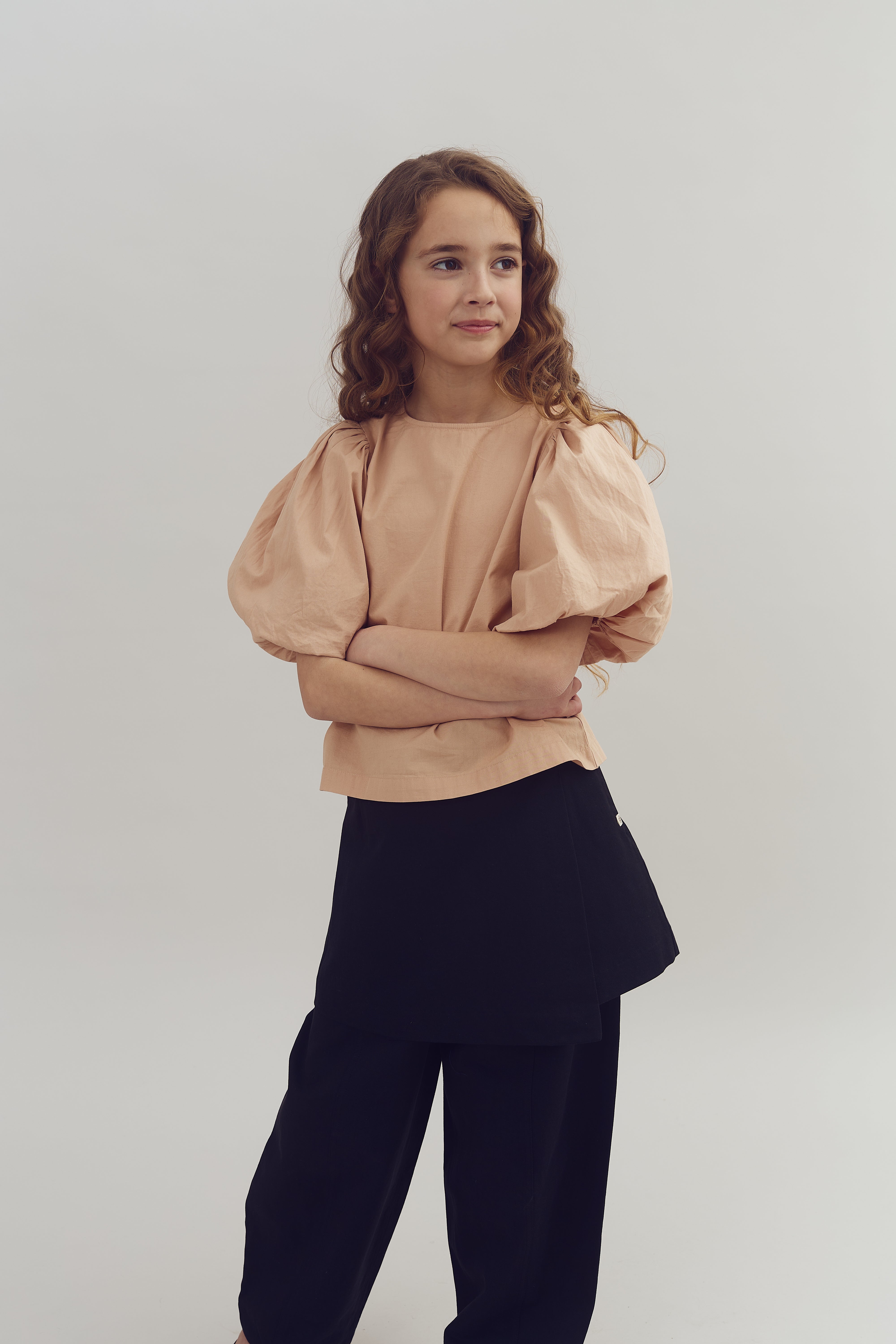 Mädchen steht mit verschränkten Armen da, trägt eine beige Bluse mit Puffärmeln und einen schwarzen Rock, vor neutralem, hellem Hintergrund.