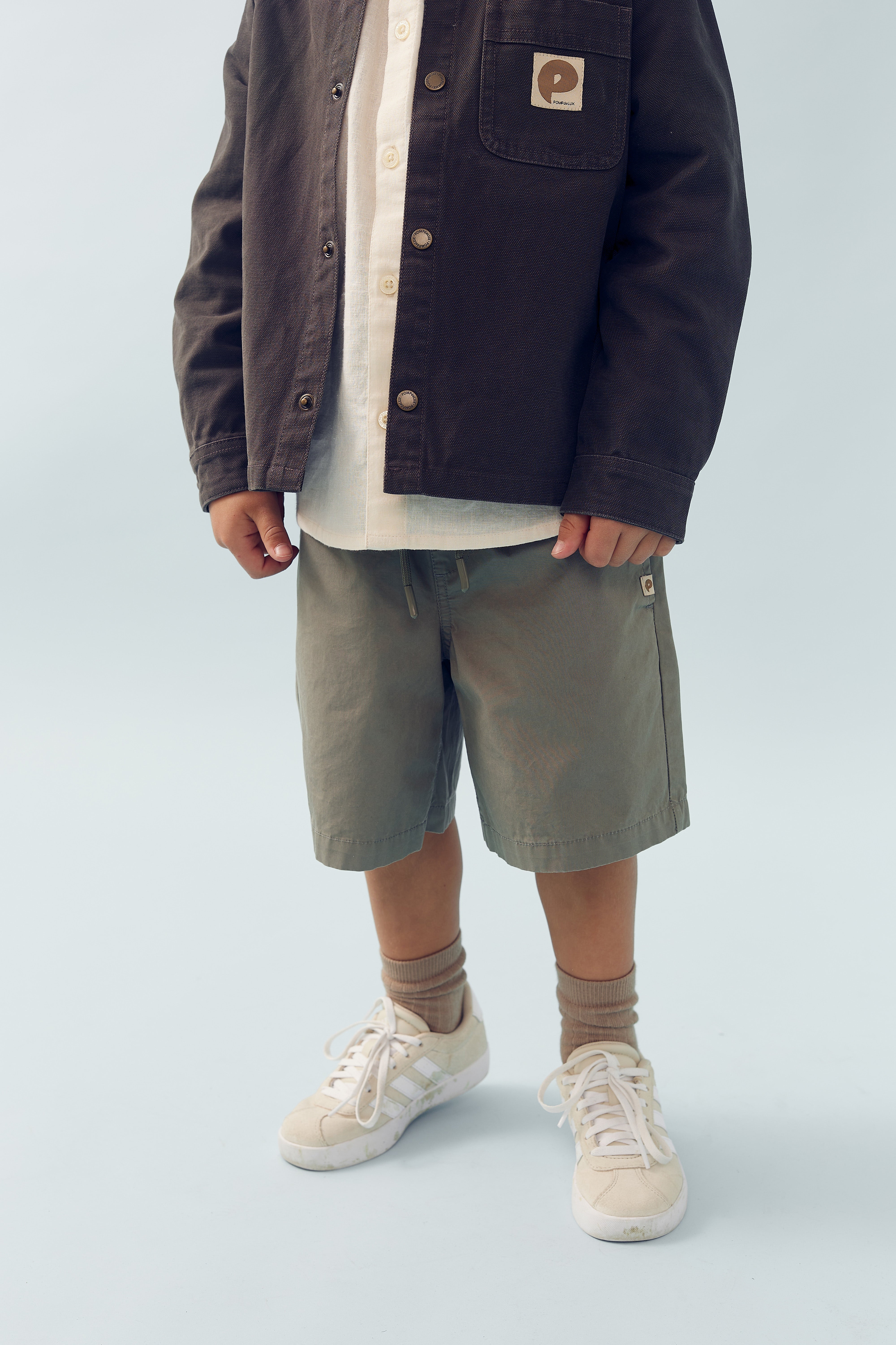Ein Kind trägt eine braune Jacke mit Logo auf der Brusttasche, ein weißes Hemd, grüne Shorts, beige Socken und weiße Turnschuhe vor einem neutralen Hintergrund.
