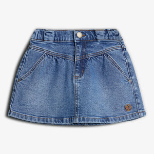 Jeansrock mit Knopfverschluss und zwei Taschen, symmetrisch angeordnet. Der Rock hat eine blaue Denim-Farbe und einen kleinen, dekorativen Aufnäher unten rechts auf dem Stoff.