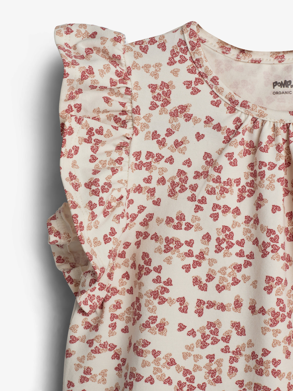 Ein weißes Kleidungsstück mit Rüschenärmeln zeigt ein Muster aus kleinen, roten und beigen Herzformen. Der Hintergrund ist neutral. Text: "POMPdeLUX ORGANIC COTTON".