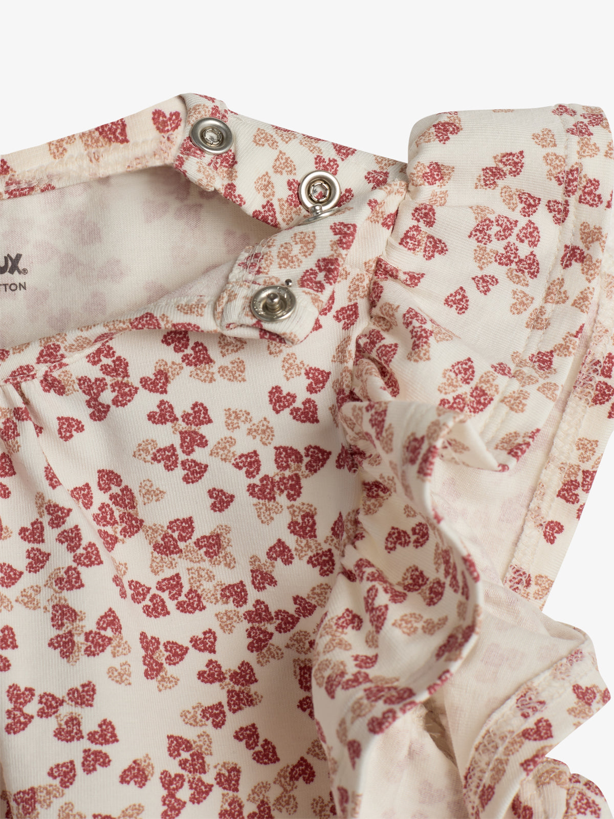 Ein Babystrampler mit rot-beigen, blumigen Stoffmuster, versehen mit Druckknöpfen an der Schulterpartie, liegt in einer hellen Umgebung. Auf dem Etikett steht "TOPOLOUX - 100% COTTON".