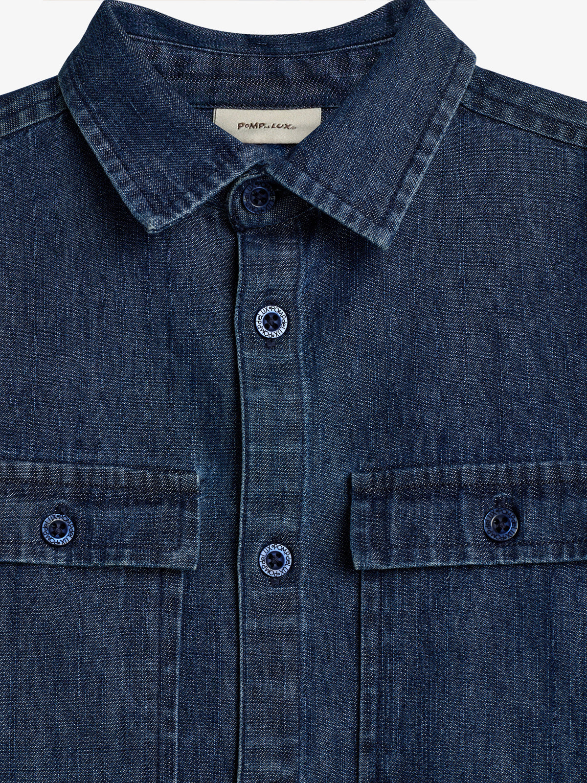 Jeansstoffhemd mit Kragen, Knopfleiste und zwei Brusttaschen. Etikett oberhalb des Kragens zeigt Text: "pomp de lux". Knöpfe tragen die Aufschrift: "U TOPIC UTOPIC." Farbtöne in dunkelblauem Denim.