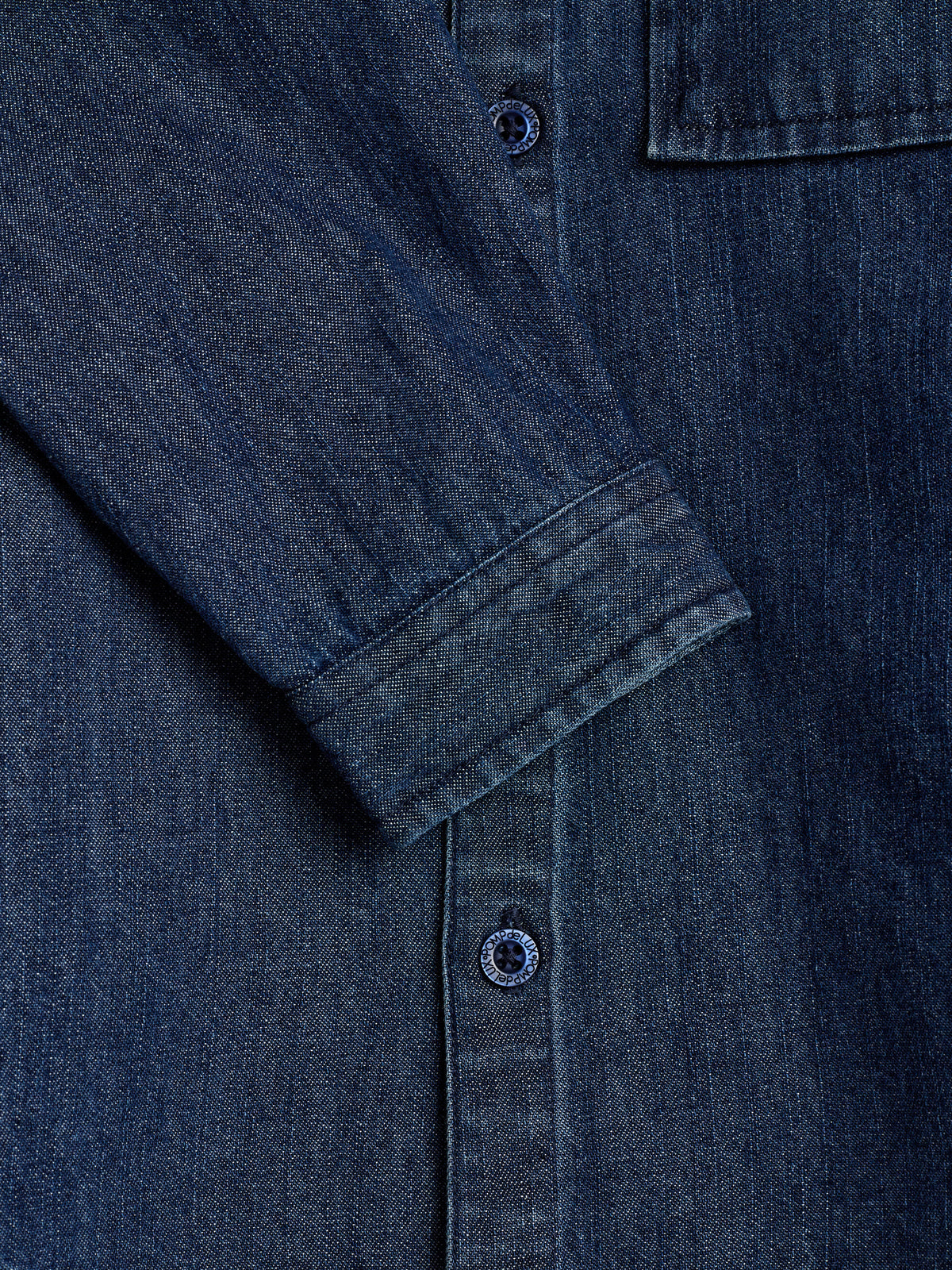 Dunkelblaue Jeansjacke mit Nahansicht eines Arms, der nach unten zeigt, und sichtbaren Knöpfen mit der Aufschrift "Kooples Club." Kontext im Bild ist ein strukturierter Stoffhintergrund.