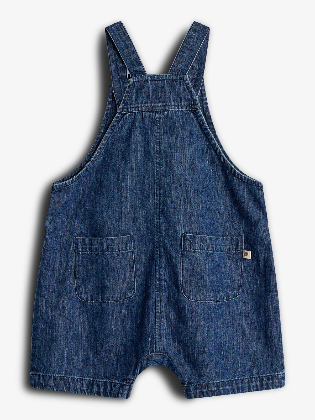 Blauer Jeans-Jumpsuit mit zwei Taschen vorne, breiten Trägern und Schürzenausschnitt. Kleine braune Stoffetikett an der Tasche. Vor weißem Hintergrund präsentiert.