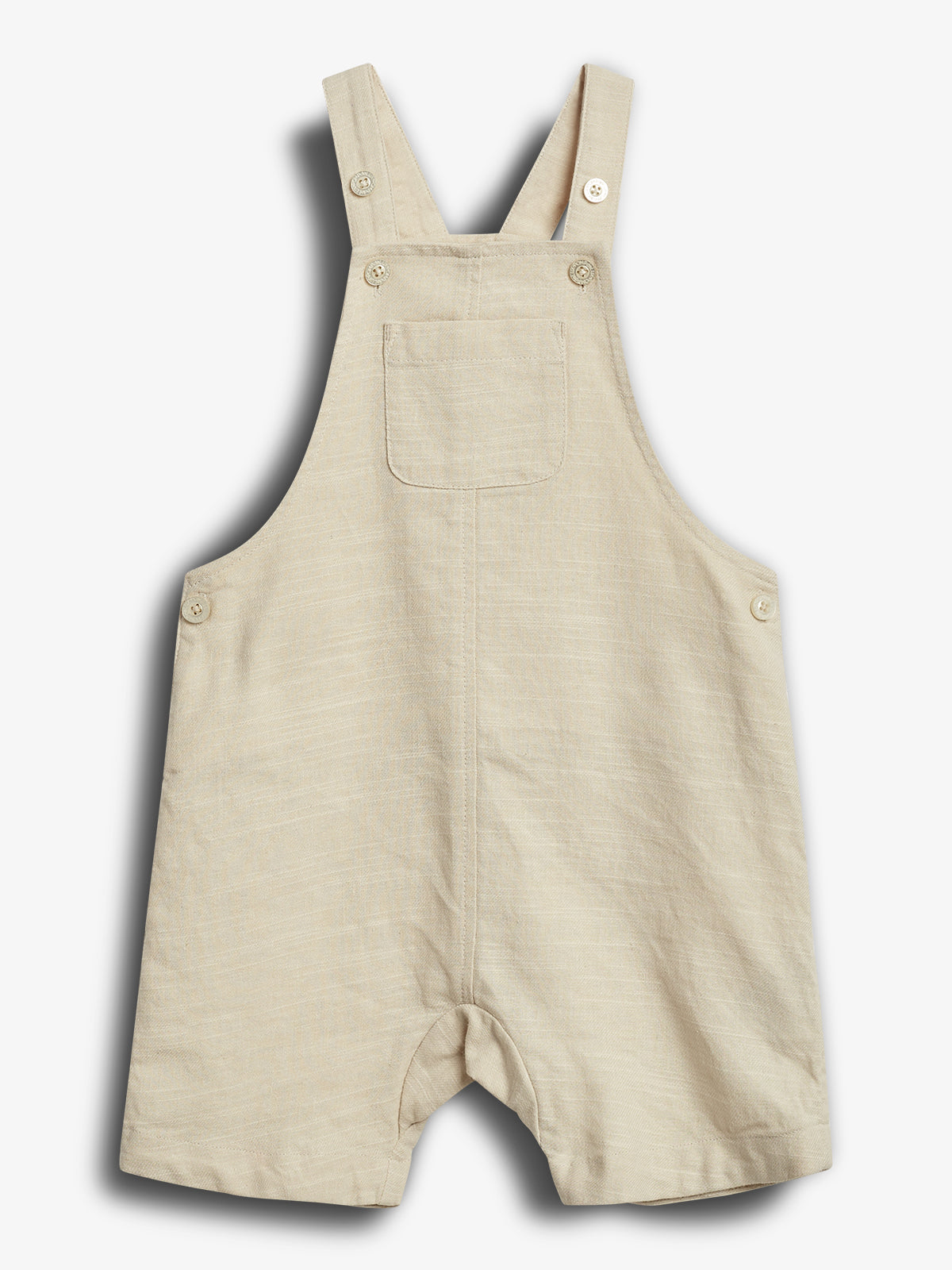 Eine cremefarbene Latzhose aus Stoff mit einem Brusttasche und Knöpfen an Trägern und Seiten. Sie liegt auf einem weißen Hintergrund.