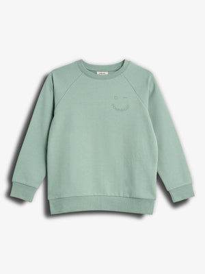Ein hellgrüner Pullover liegt flach vor einem weißen Hintergrund. Oben rechts ist ein dezentes Gesichtsmotiv mit dem Text "Bonjour." sichtbar.