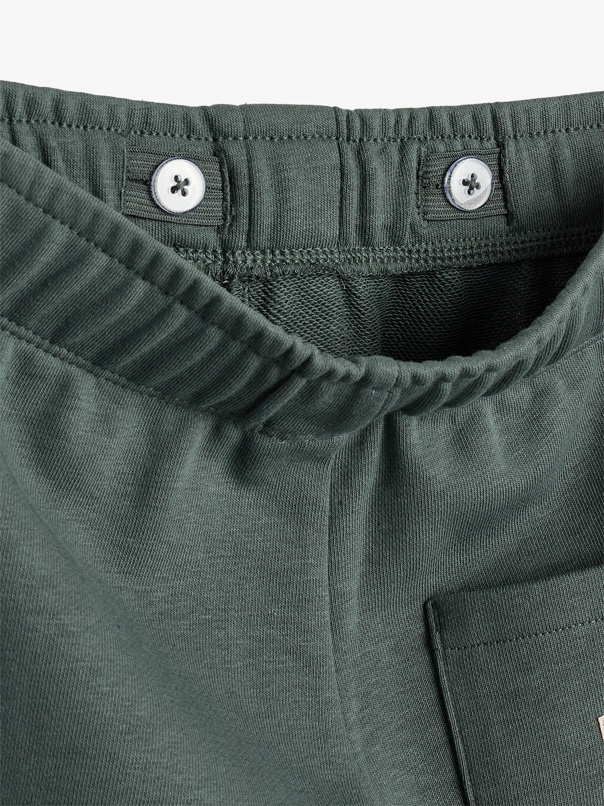 Ein Bund einer dunkelgrünen Hose zeigt zwei weiße Knöpfe zur Anpassung. Die Tasche ist teilweise sichtbar. Der Hintergrund ist neutral, was den Stoff betont.