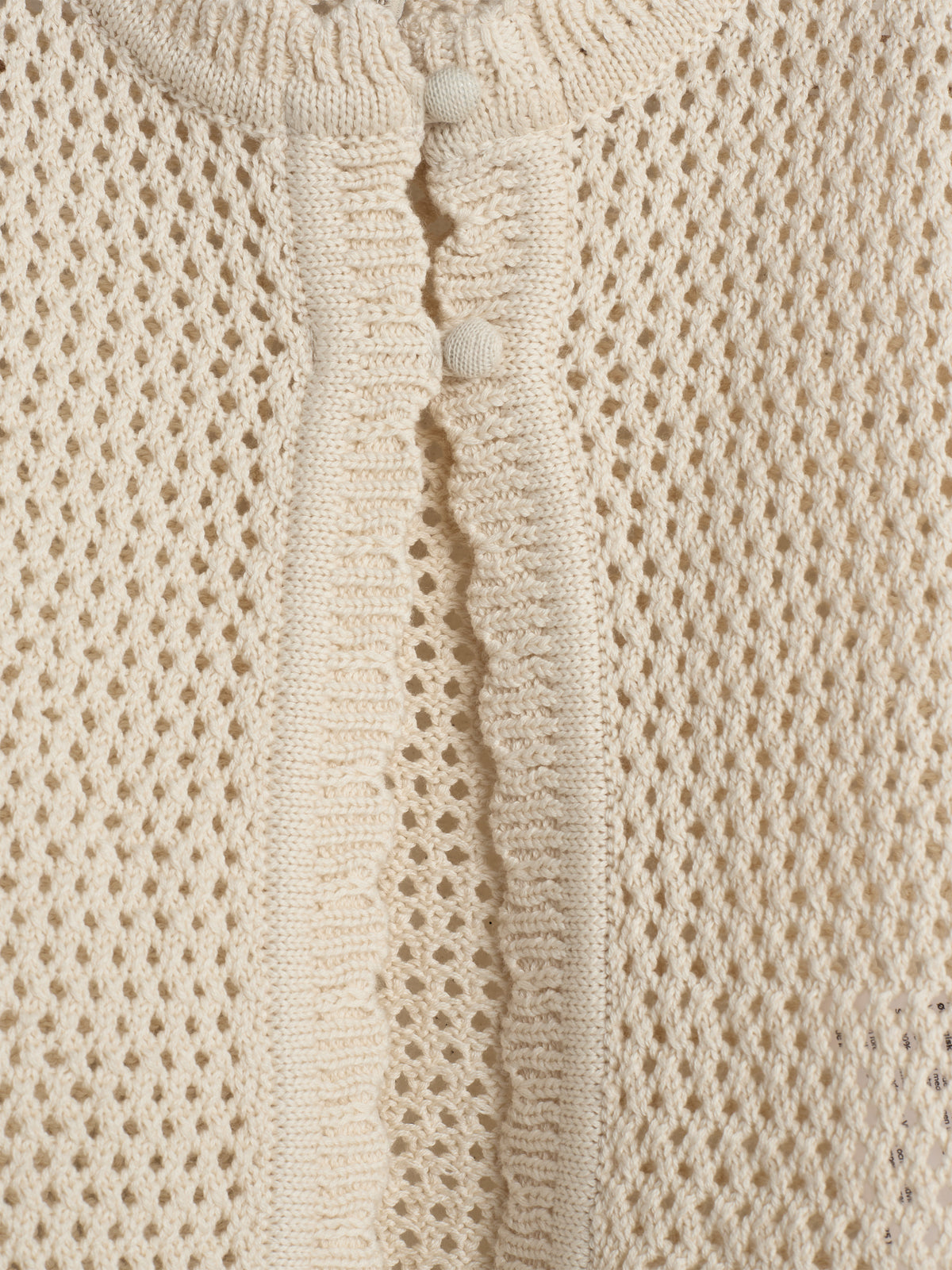 Beigefarbener Strickstoff mit Lochmuster, an dem zwei kleine Knöpfe angenäht sind. Das Muster wiederholt sich gleichmäßig, was dem Material ein texturiertes Aussehen verleiht.