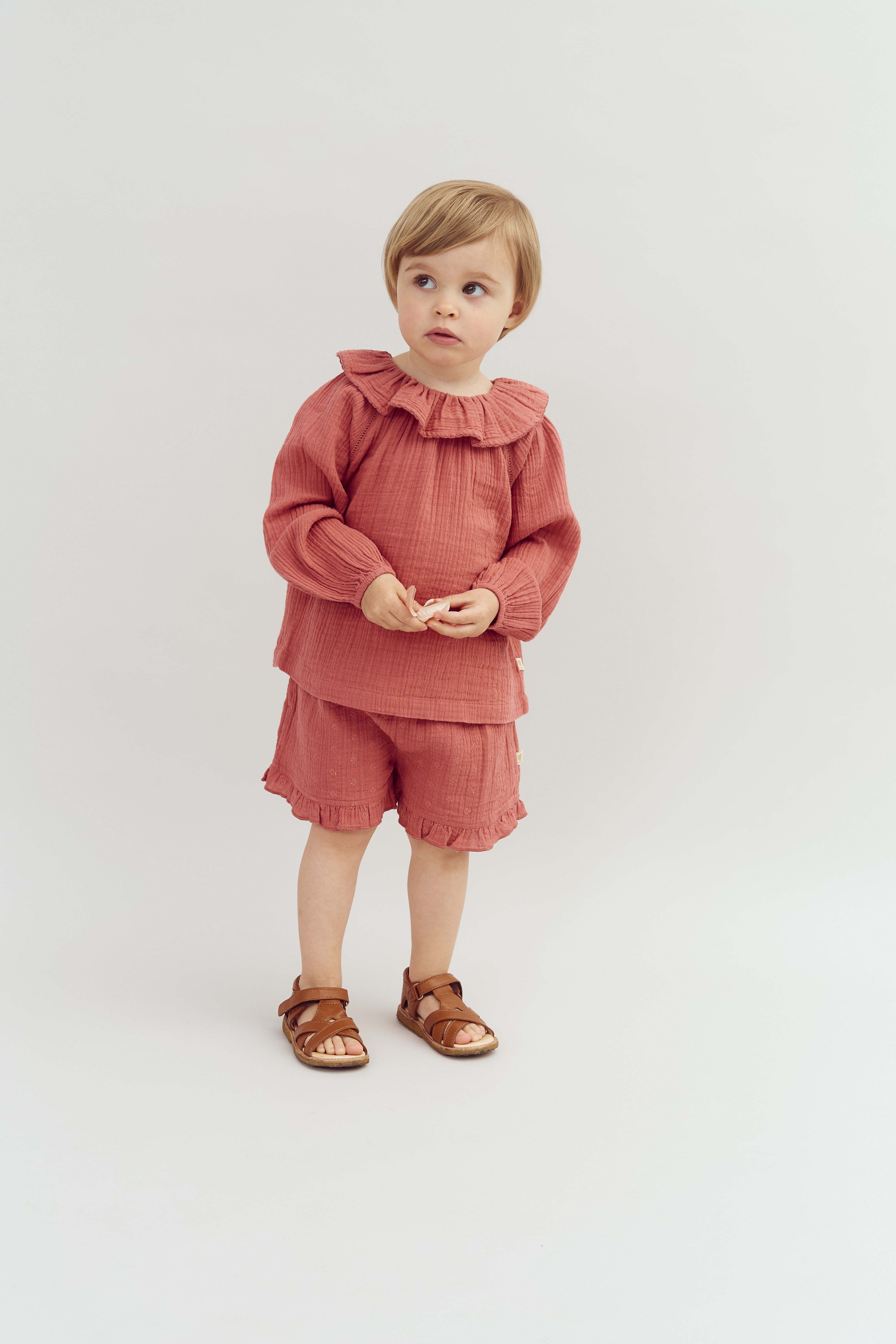 A child in a pink ruffled outfit stands on a white background, looking off to the side with hands together, wearing brown sandals.