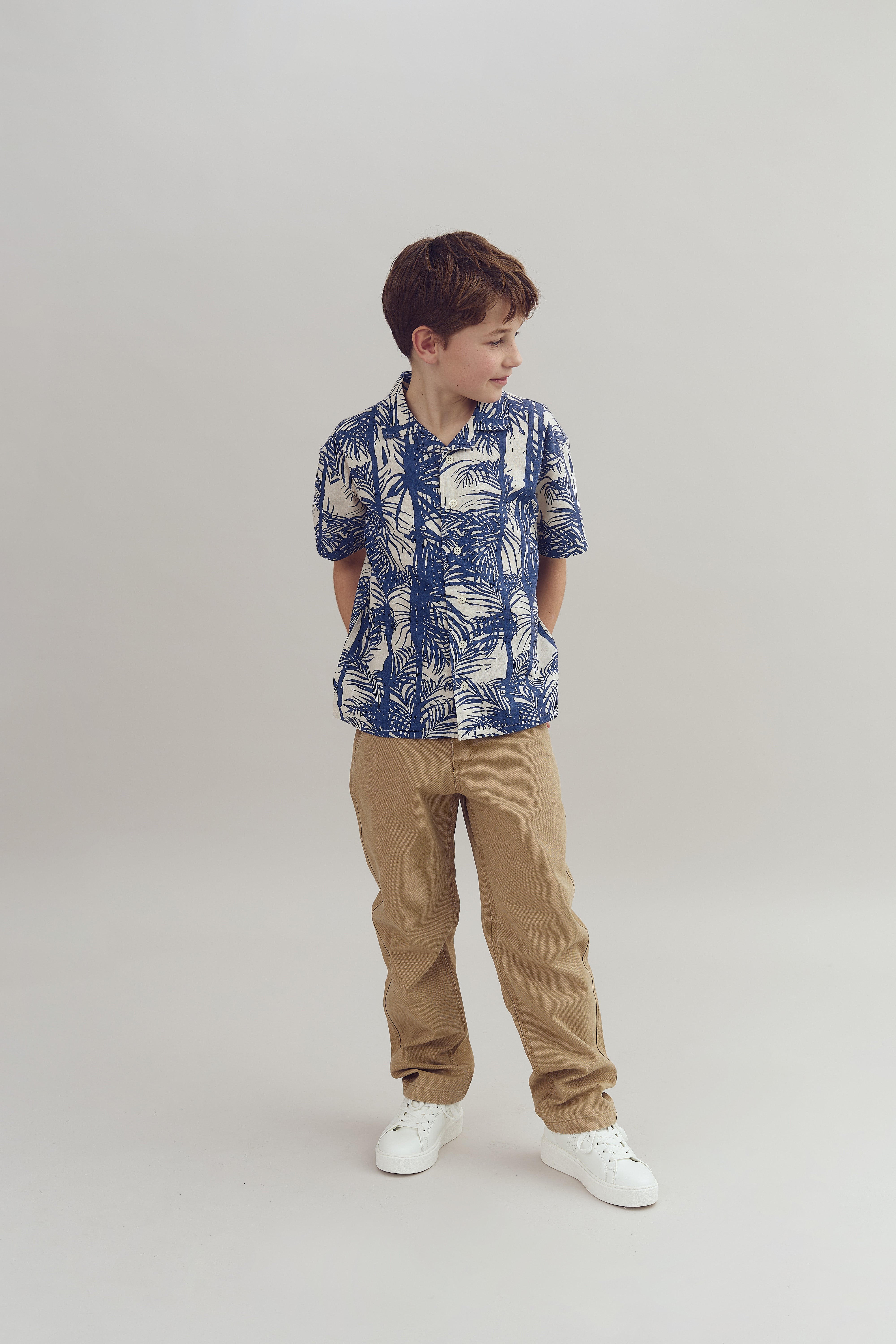 Ein Junge steht mit den Händen hinter dem Rücken. Er trägt ein blau-weißes Hawaiihemd und beige Hosen. Der Hintergrund ist neutral und schlicht.