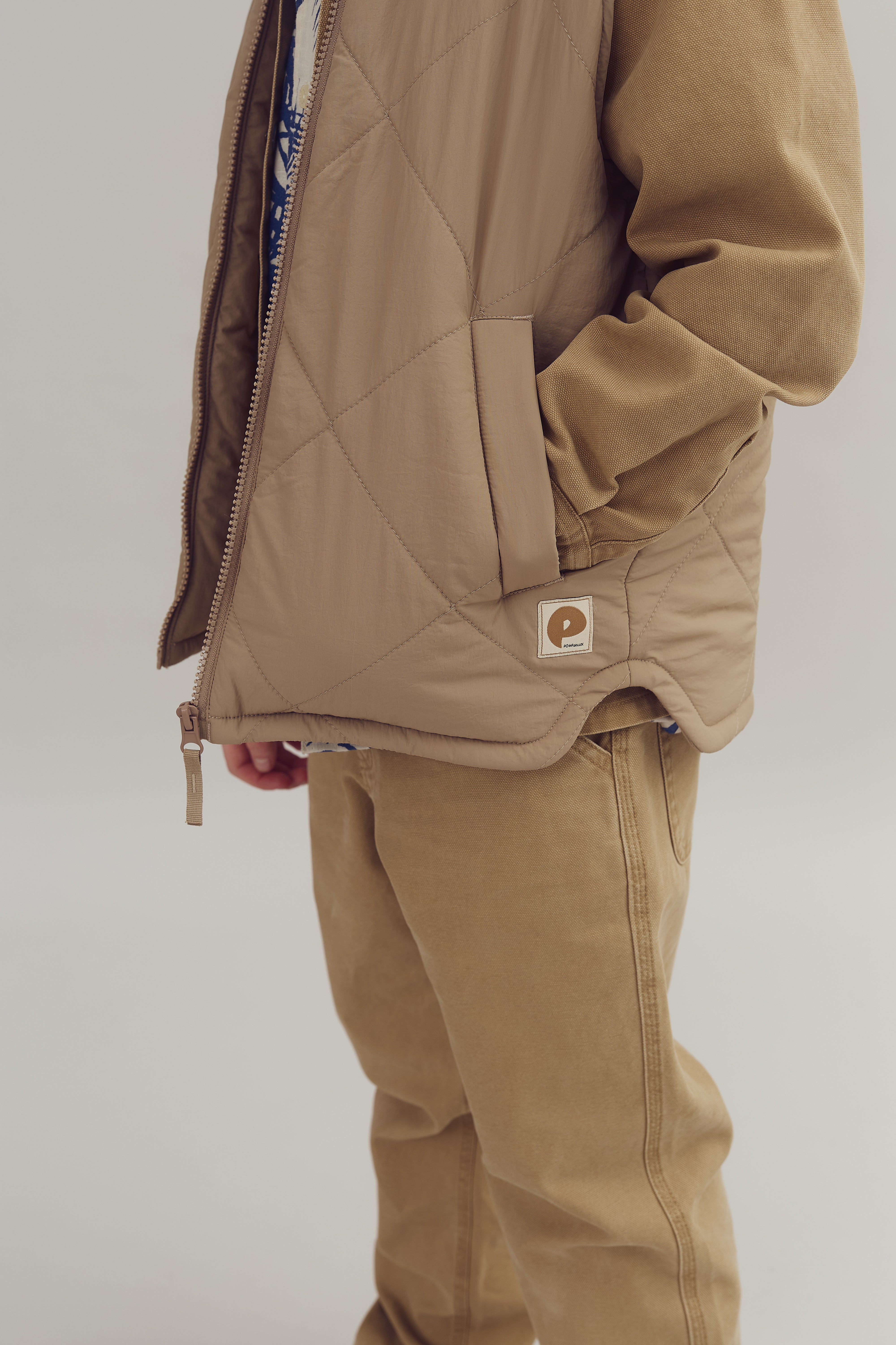Jacke in Beige mit gestepptem Muster und sichtbarem Reißverschluss. Person trägt die Hände in den Taschen, Jeans in ähnlicher Farbe. Neutraler Hintergrund. Patch mit Schriftzug "Powers" sichtbar.