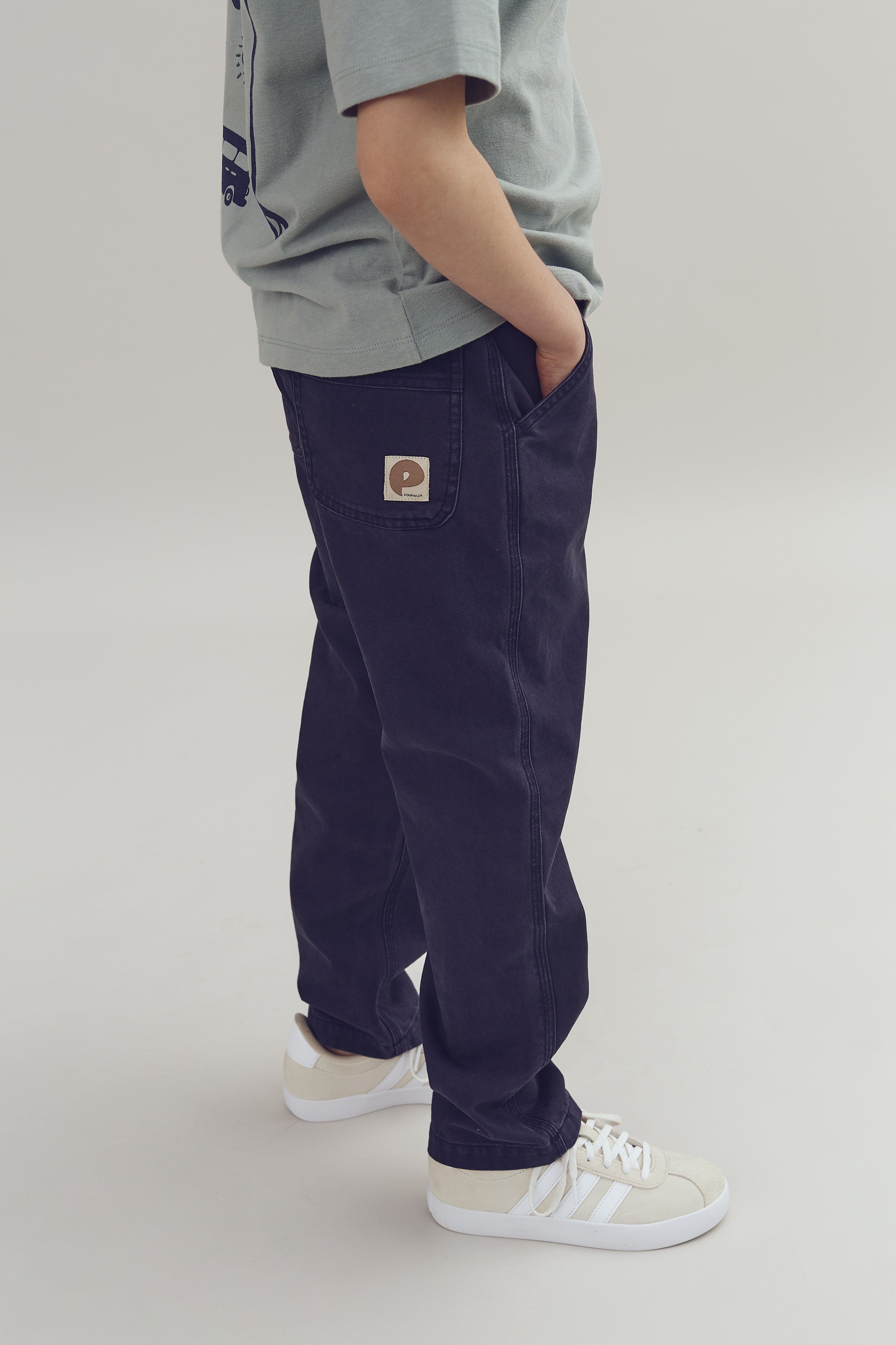 Ein Kind trägt eine graue Hose mit einem aufgenähten Etikett, die Hände in den Taschen. Es ist in einem grauen Raum, trägt ein graues T-Shirt und weiße Turnschuhe.