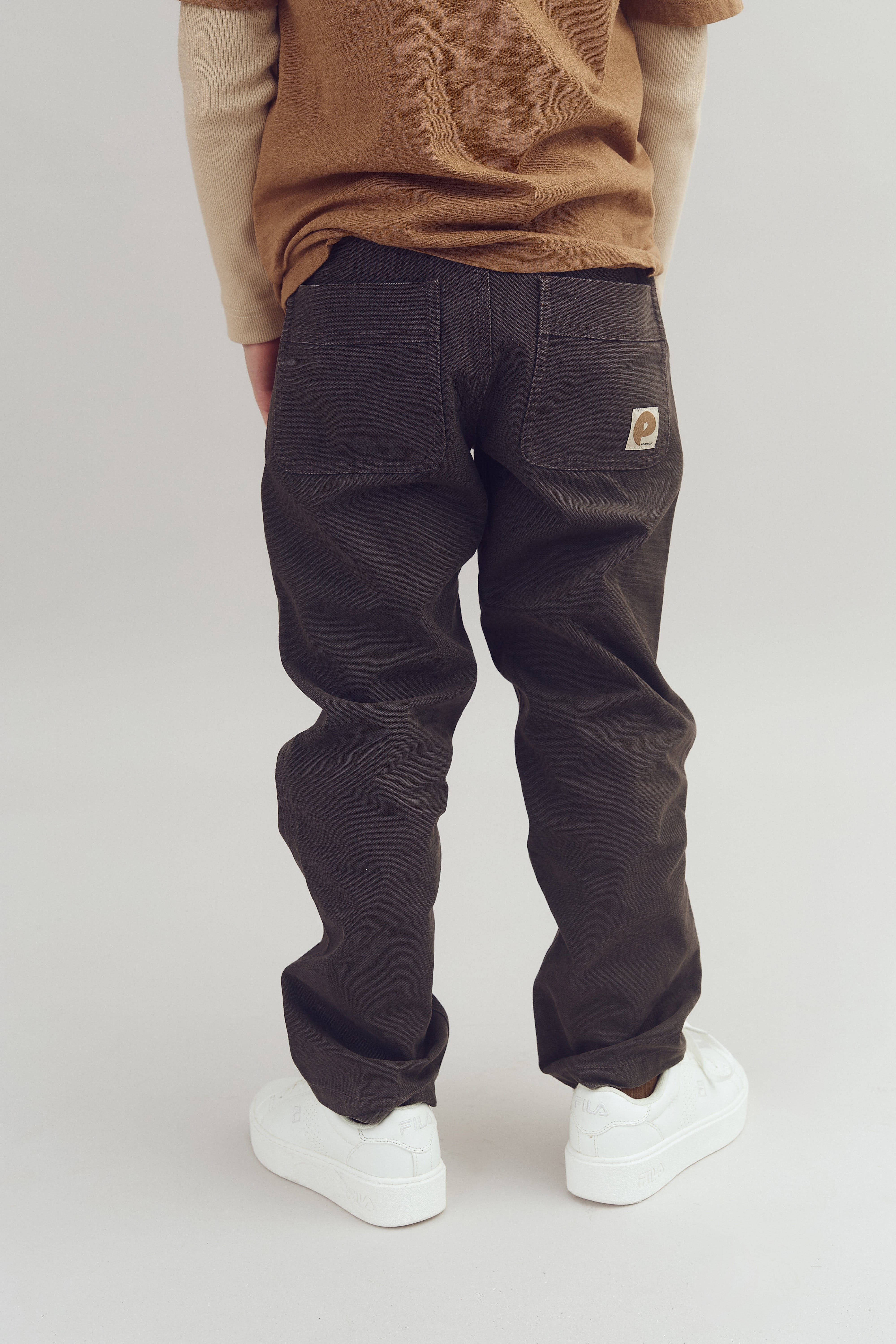 Eine Person steht mit dem Rücken zur Kamera. Sie trägt dunkle Hose mit zwei Taschen und ein braunes Oberteil. Weiße Schuhe mit der Aufschrift "FILA" befinden sich auf einem hellen Hintergrund.