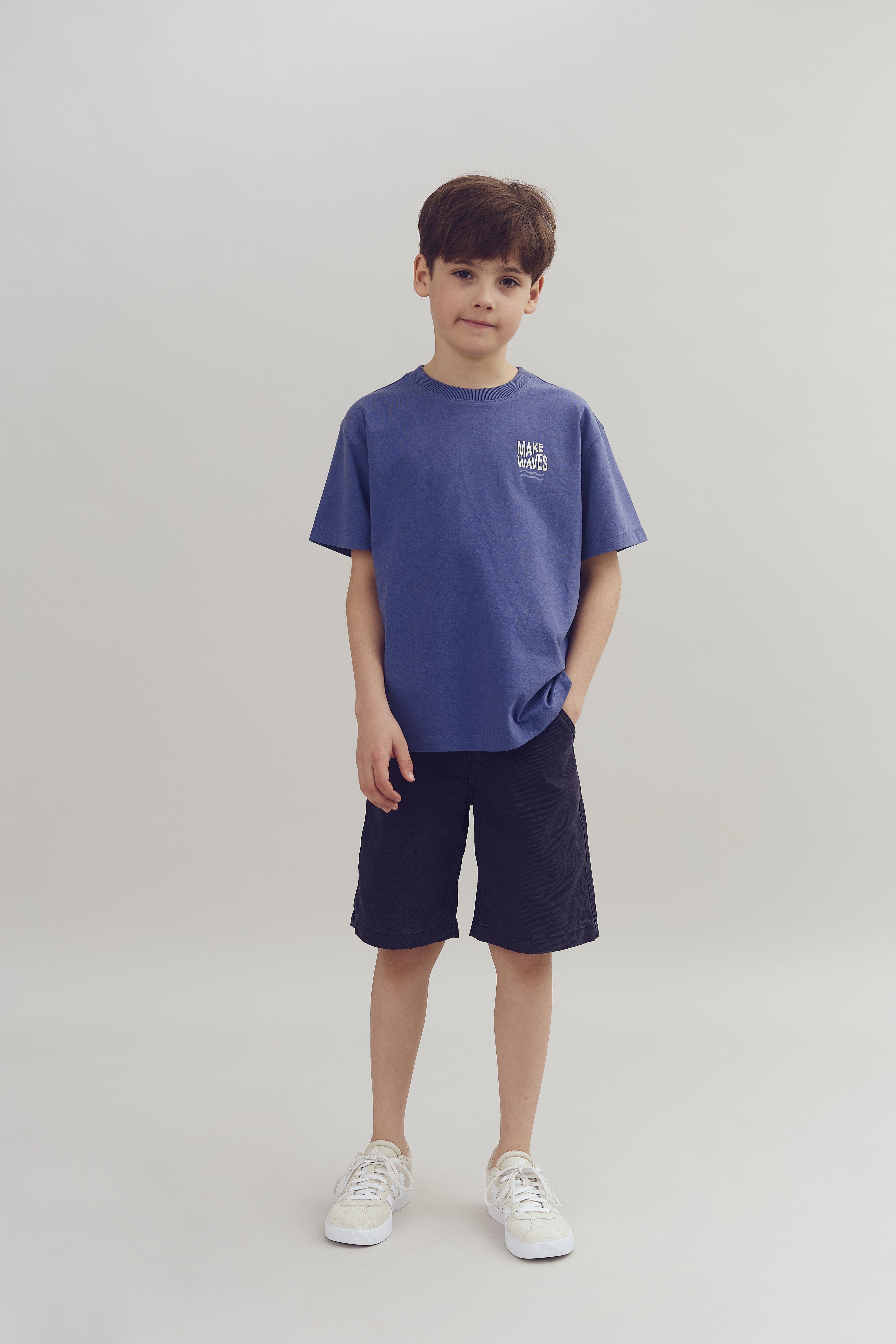 Ein Junge steht in einem neutralen Raum. Er trägt ein blaues T-Shirt mit der Aufschrift "MAKE WAVES", dunkle Shorts und weiße Turnschuhe.
