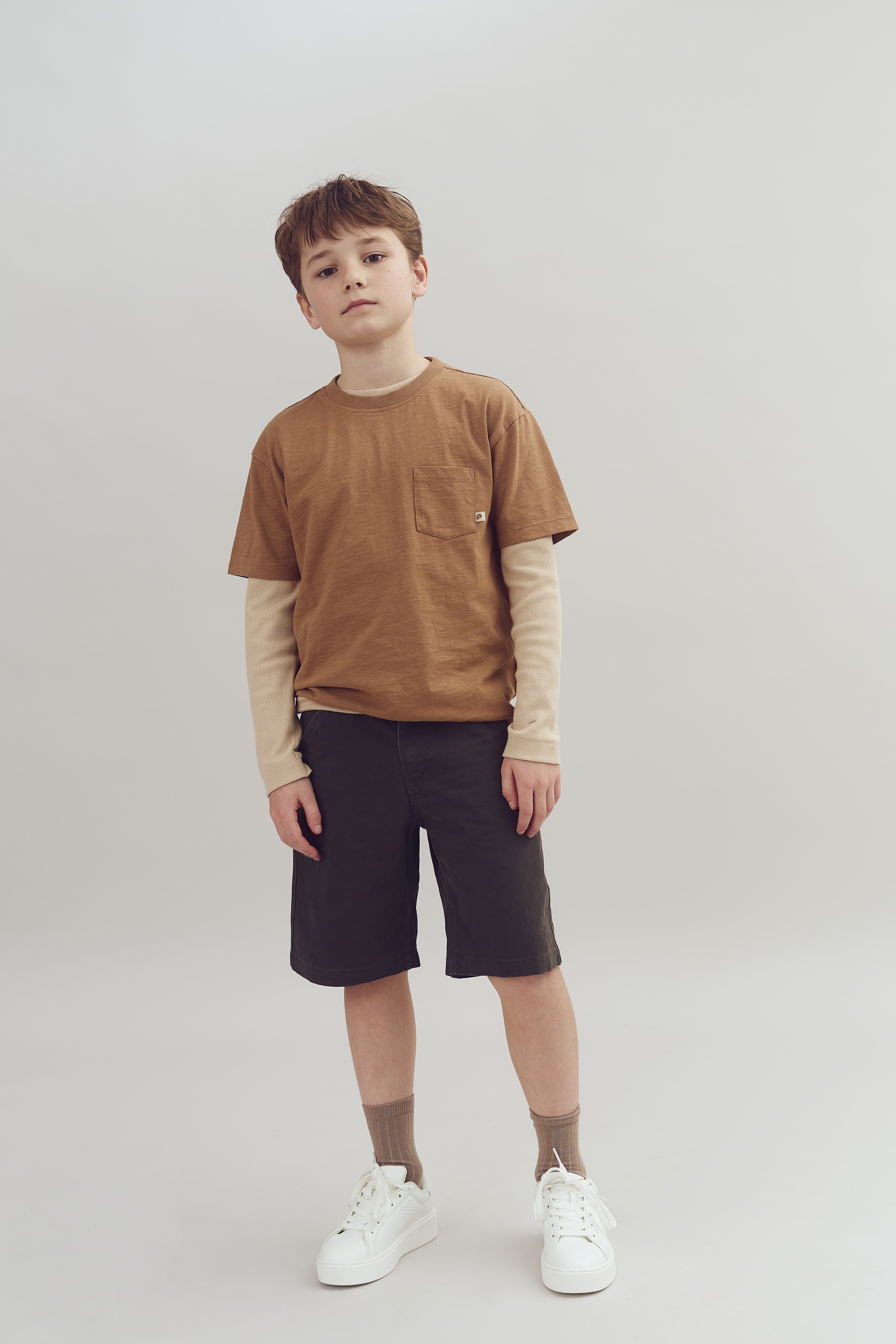 Ein Junge steht ruhig, trägt ein braunes T-Shirt über einem langen cremefarbenen Shirt, dunkle Shorts und weiße Sneakers, vor einem neutralen Hintergrund.
