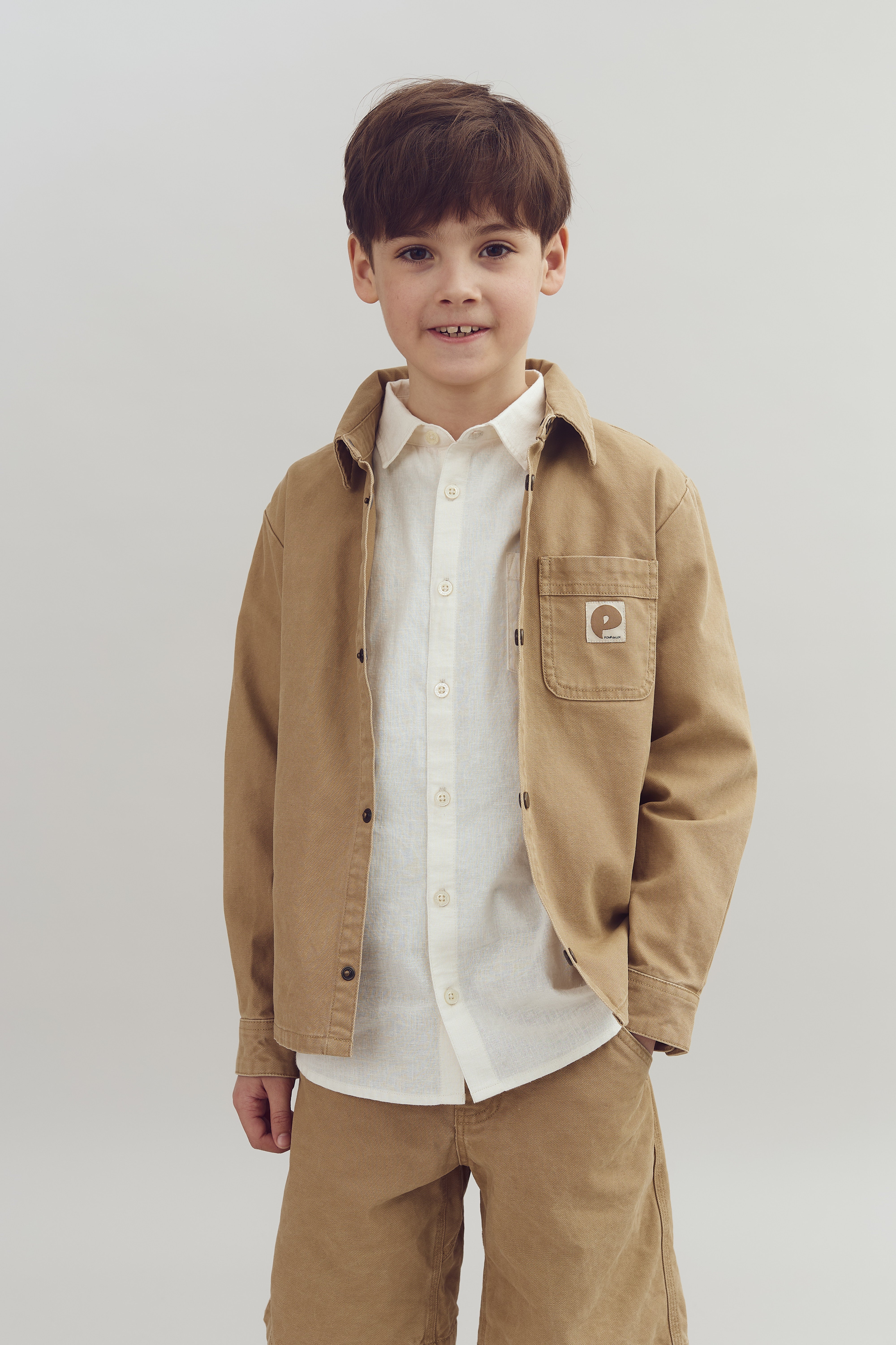 Junge lächelt in Kamera, trägt helles Hemd und beige Jacke mit Logo „popelin“ auf Tasche, steht vor neutralem, grauem Hintergrund.