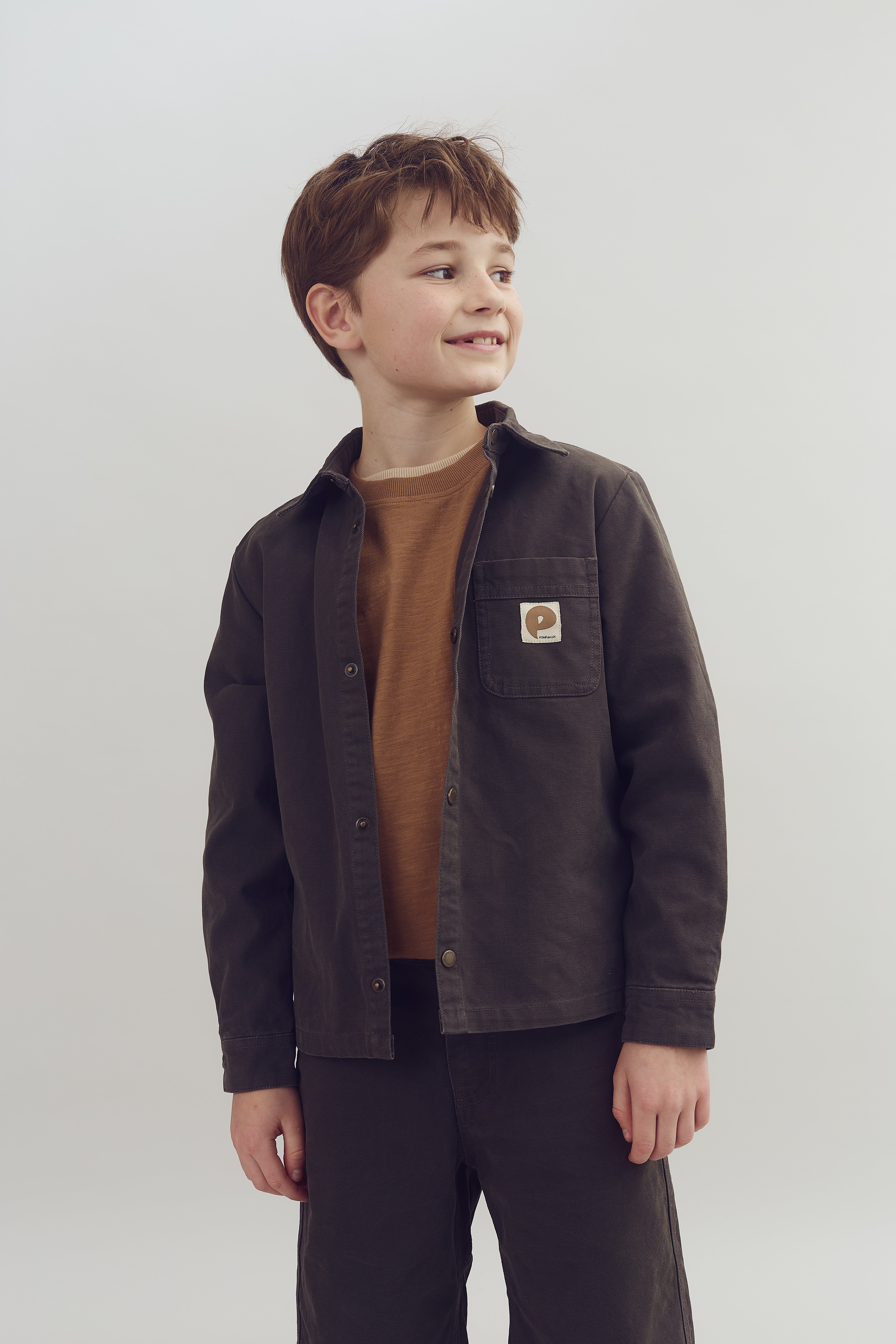 Ein Junge trägt eine braune Jacke und schaut lächelnd nach rechts. Ein Emblem mit einem "p" ist auf der Brusttasche. Der Hintergrund ist hell und neutral.