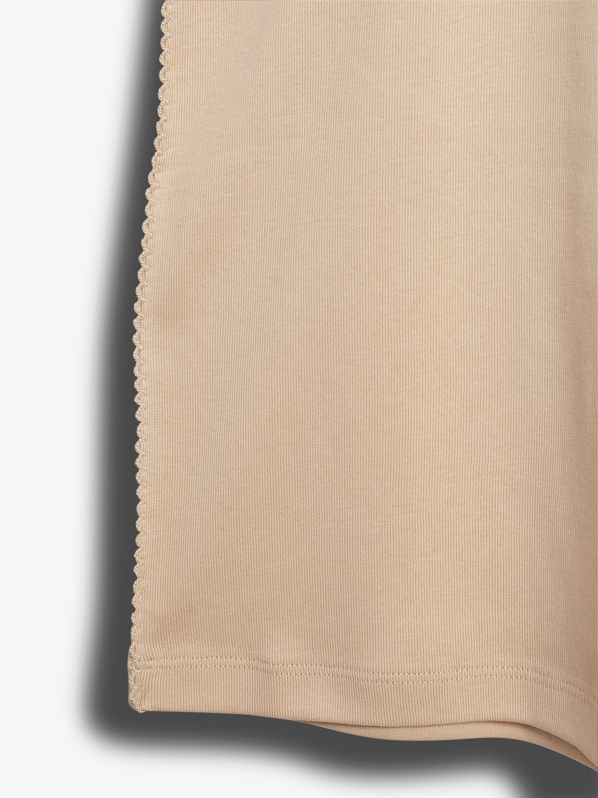 Beige, seidiges Kleidungsstück hängt mit einer dekorativen Kante im Tortenstil an der Seite. Der Stoff wirft einen Schatten auf einen weißen Hintergrund.