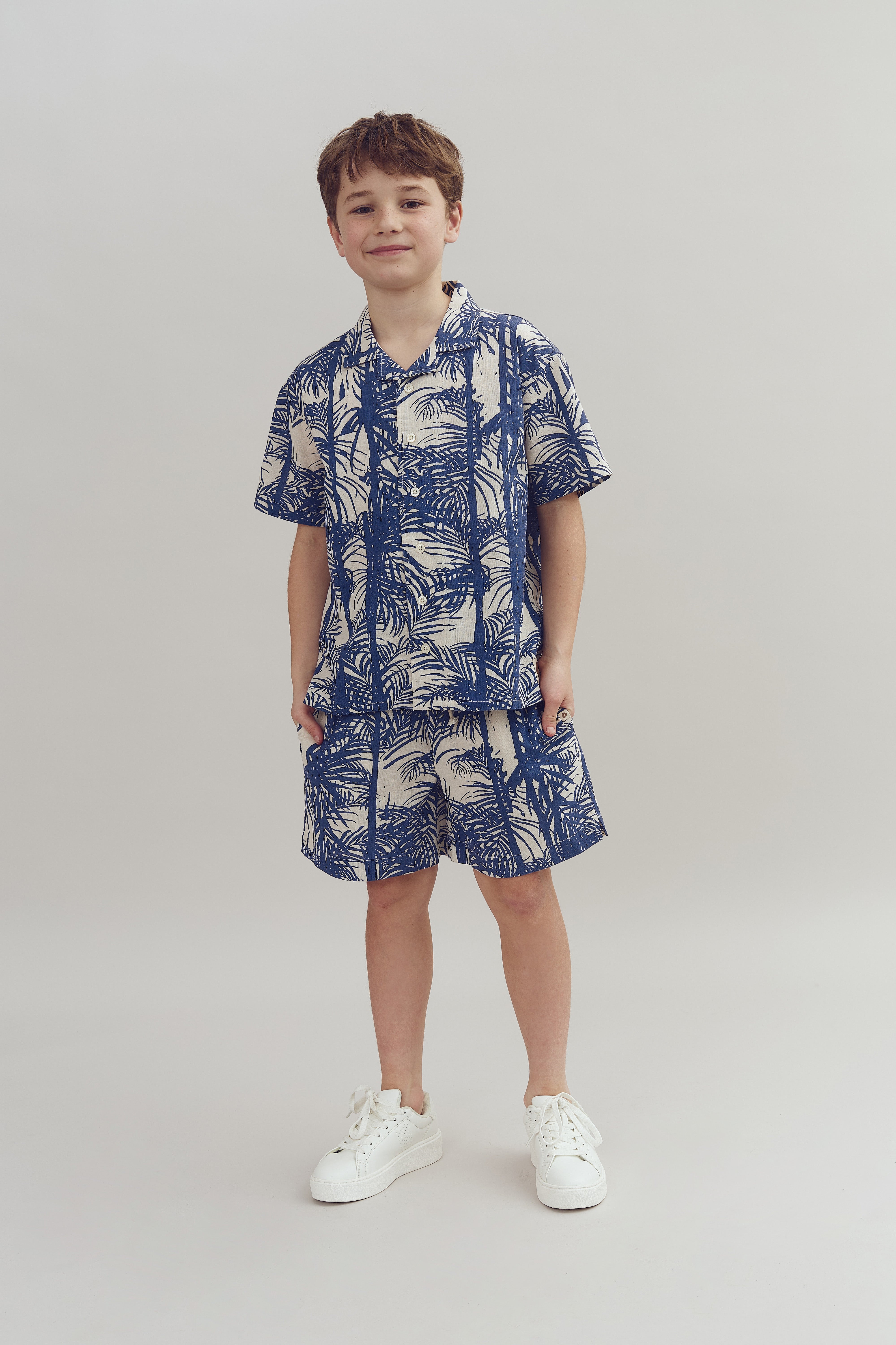 Ein Junge trägt ein blau-weiß gemustertes Hemd und Shorts, steht selbstbewusst und lächelt leicht in einer neutralen, hellen Umgebung. Er hat weiße Turnschuhe an.