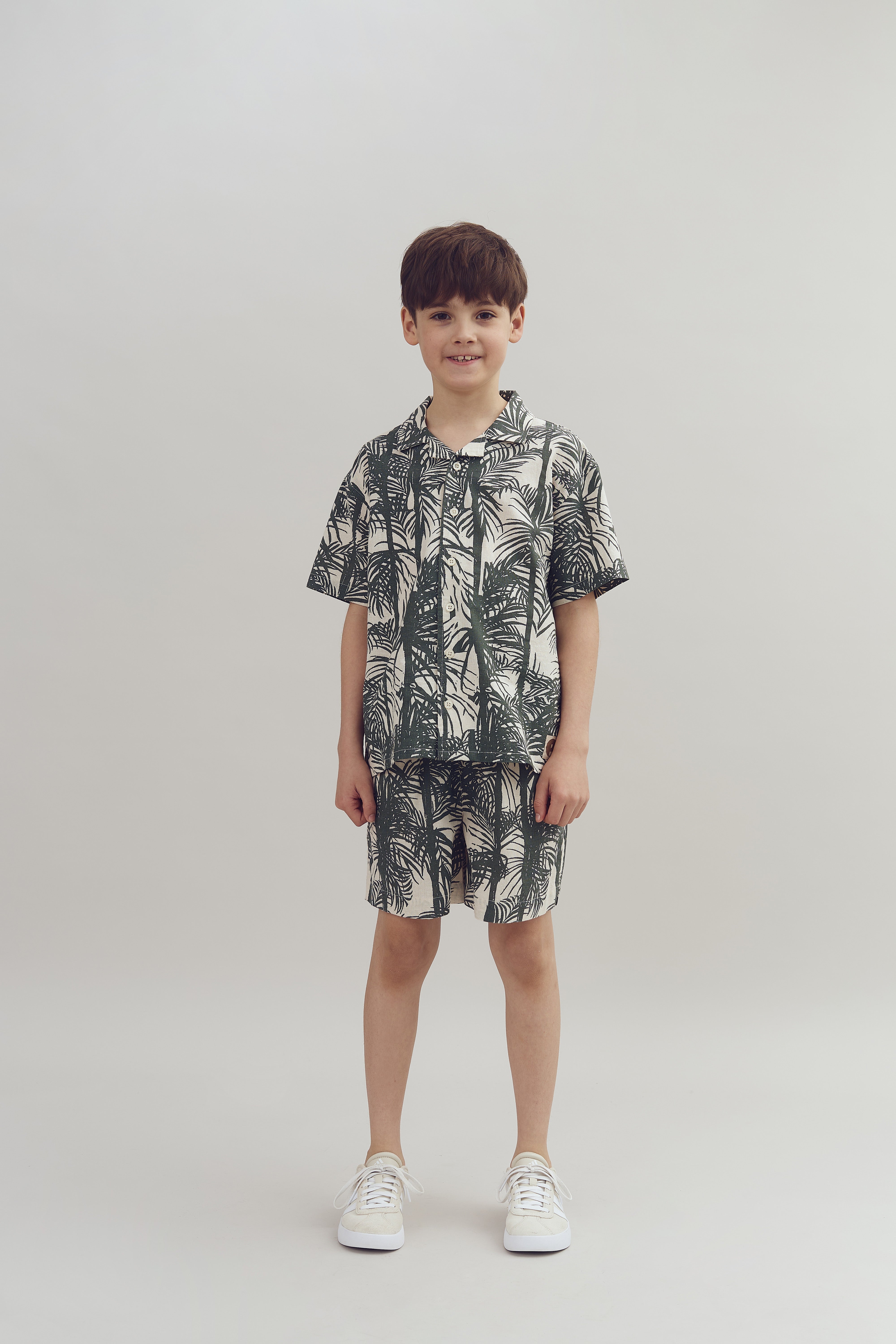 Ein Junge trägt eine kurze, tropisch gemusterte, beige und dunkelgrüne Sommercombo bestehend aus Hemd und Shorts, stehend auf grauem Studiohintergrund, und lächelt leicht.