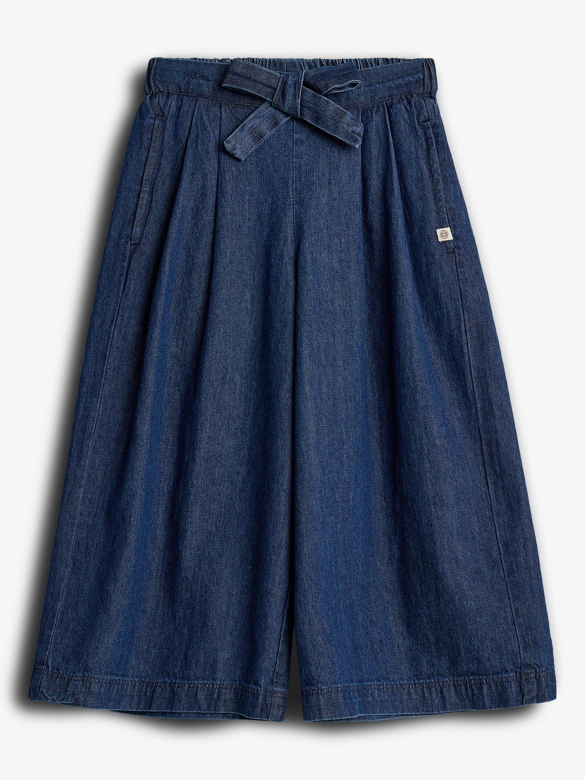 Eine dunkelblaue, weit geschnittene Culotte mit Bindegürtel liegt flach auf hellem Hintergrund. Zwei seitliche Taschen und ein kleines quadratisches Label ergänzen das schlichte Design.