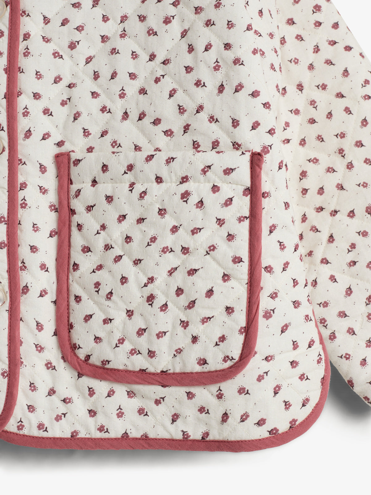 Eine gesteppte Jacke mit einem kleinen roten Blumenmuster bedeckt die Oberfläche. Sie hat eine rote Paspelierung und eine aufgesetzte Tasche. Der Hintergrund ist neutral.