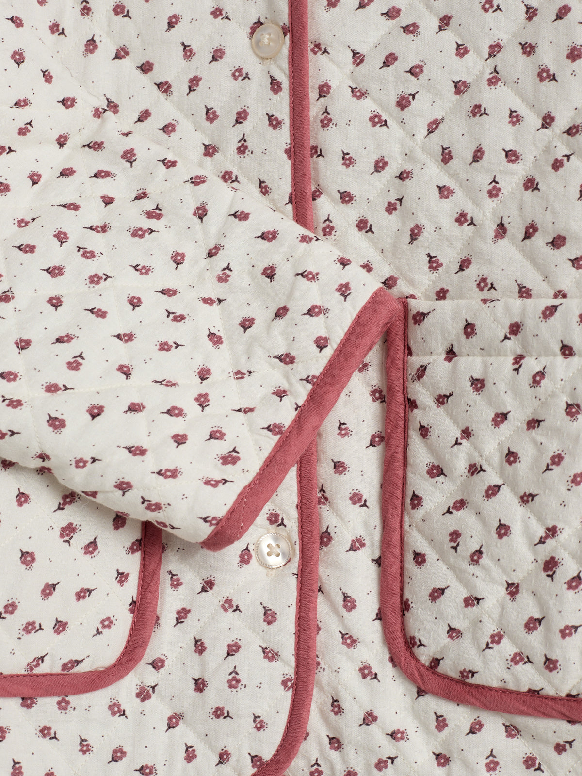 Eine gestepptes Kleidungsstück mit rosa Blumenmuster, rotem Saum und Knöpfen. Im Vordergrund ist eine Tasche erkennbar. Es liegt auf einer glatten Oberfläche.