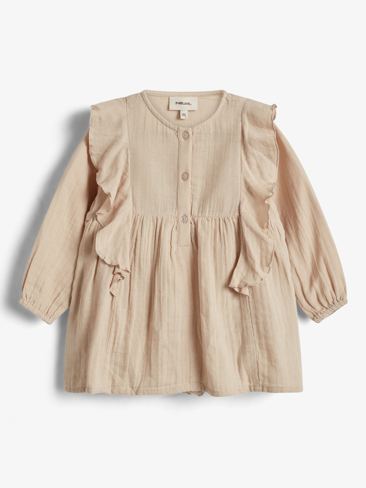 Ein beigefarbenes Kinderkleid mit langen Ärmeln und Rüschen, Knopfleiste vorne, hängt auf weißem Hintergrund. Das Etikett zeigt "PAPU 86".