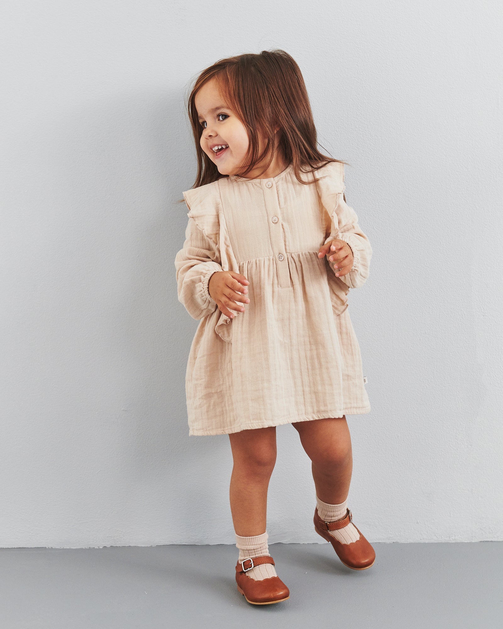 Ein kleines Mädchen lächelt und trägt ein beigefarbenes Kleid. Sie steht auf einem grauen Boden vor einer neutralen Wand und trägt braune Schuhe mit Socken.