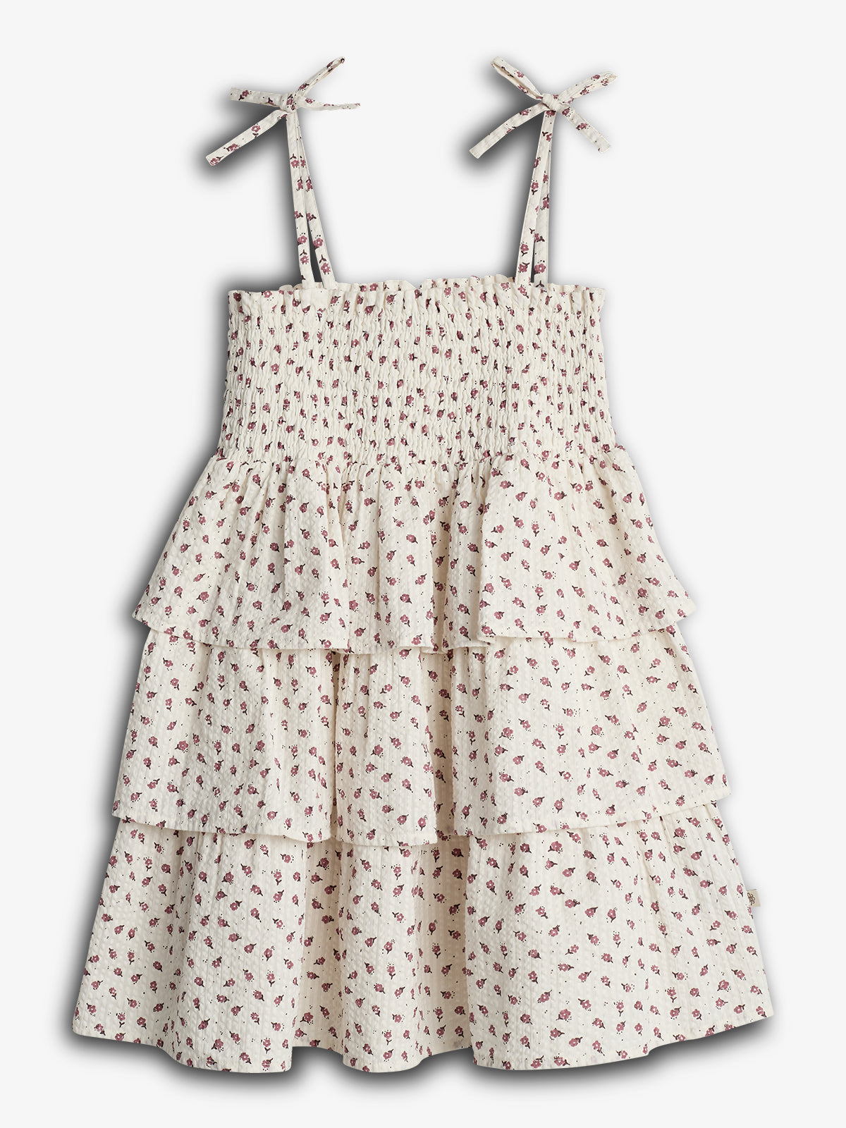 Ein ärmelloses, weißes Kleid mit kleinen, rosafarbenen Blumenmotiven und mehreren Rüschenstufen. Es hat verstellbare Träger mit Schleifen. Der Hintergrund ist neutral.