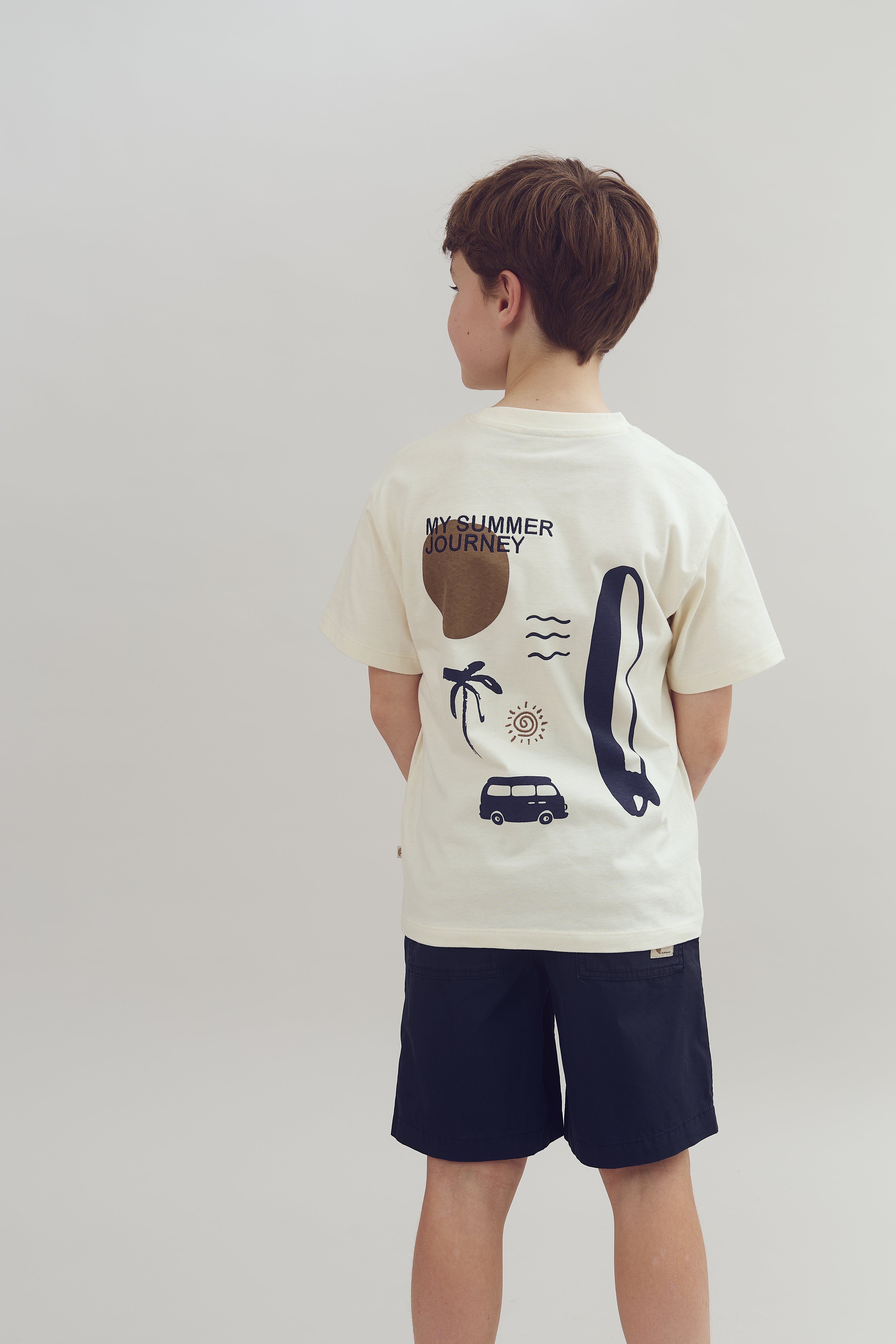 Ein Kind steht mit dem Rücken zur Kamera, trägt ein cremefarbenes T-Shirt mit der Aufschrift "MY SUMMER JOURNEY" und Sommermotiven, in einem neutralen, hellgrauen Raum.