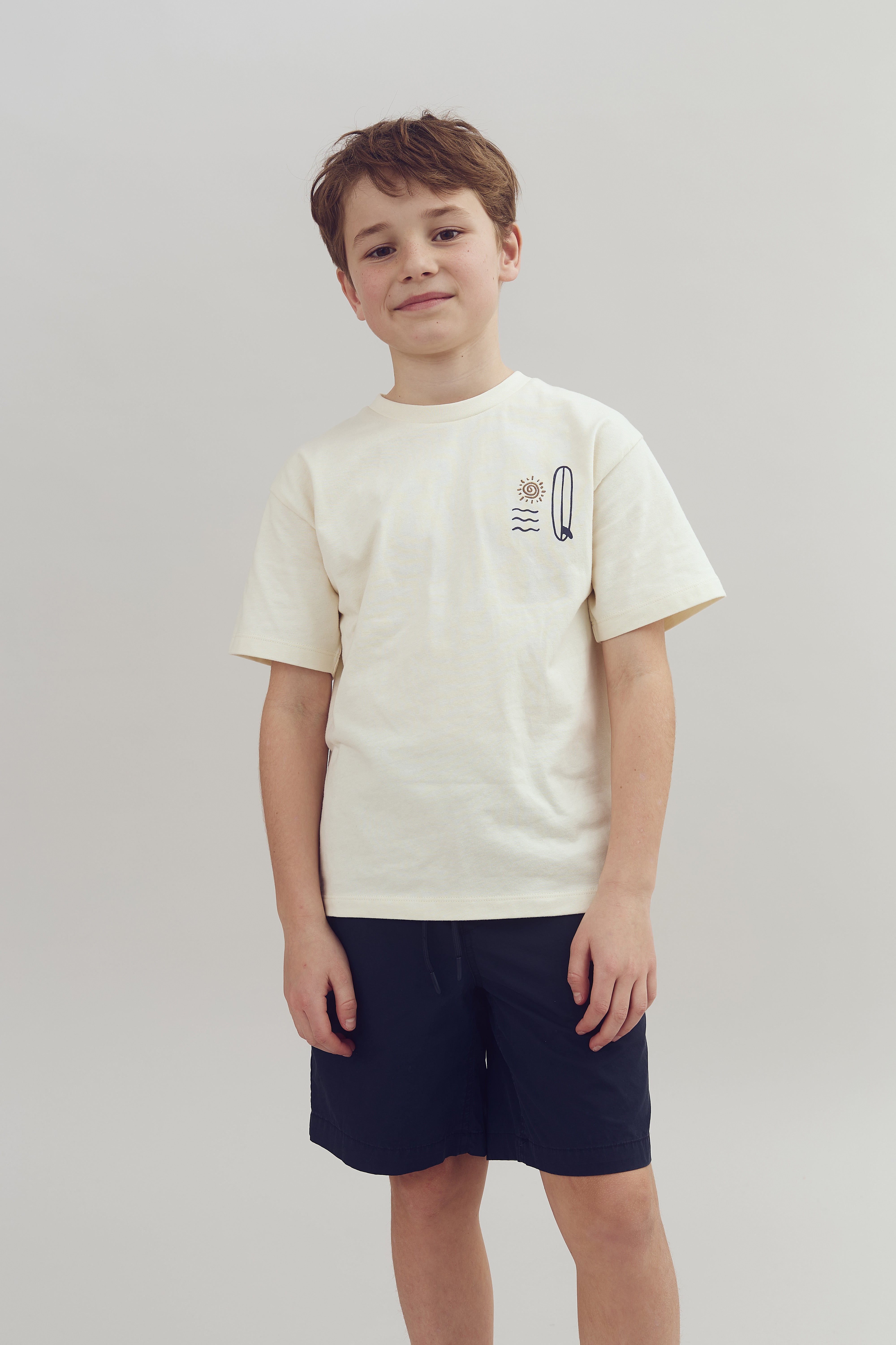 Junge steht ruhig, trägt ein cremefarbenes T-Shirt mit Sonnen- und Wellenmotiv links und dunkelblaue Shorts, neutraler Hintergrund.