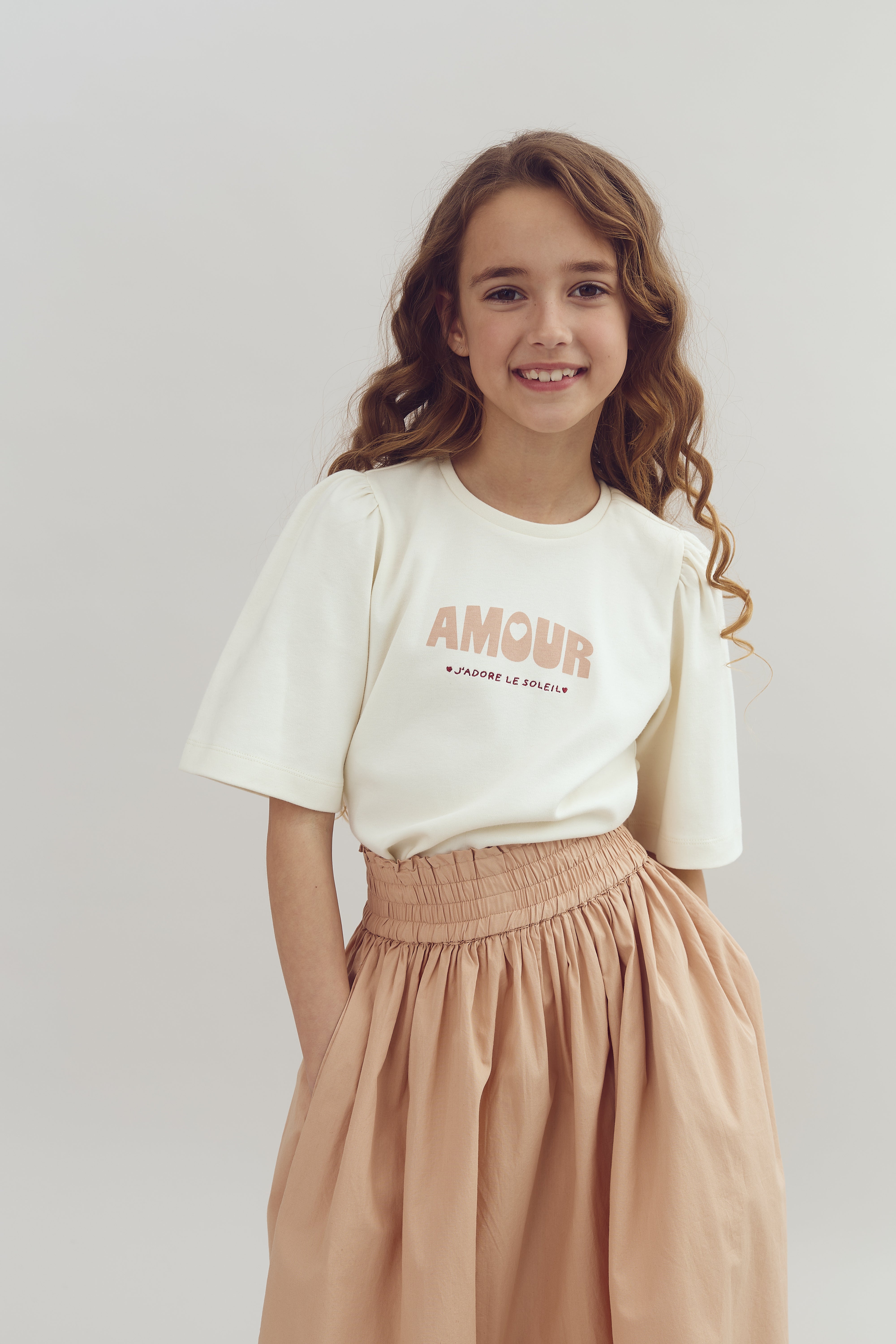 Mädchen lächelt, trägt ein weißes T-Shirt mit der Aufschrift "AMOUR: J'ADORE LE SOLEIL" und einen langen, beigen Rock. Sie steht vor einem neutralen, hellen Hintergrund.