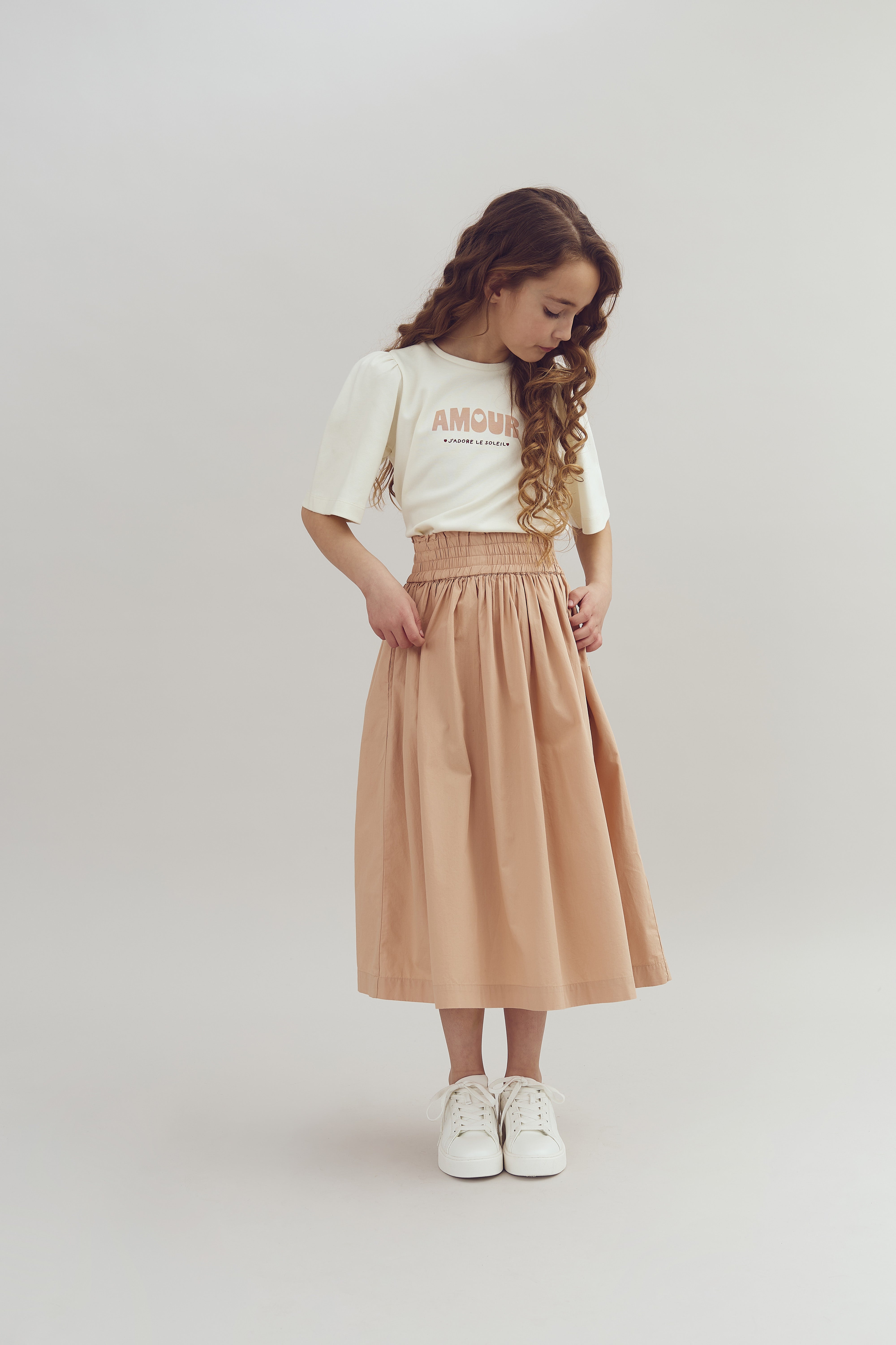 Ein Mädchen steht in einem Studio, betrachtet den Boden, trägt ein cremefarbenes T-Shirt mit der Aufschrift "AMOUR" und einen langen beigen Rock, umgeben von einem neutralen Hintergrund.