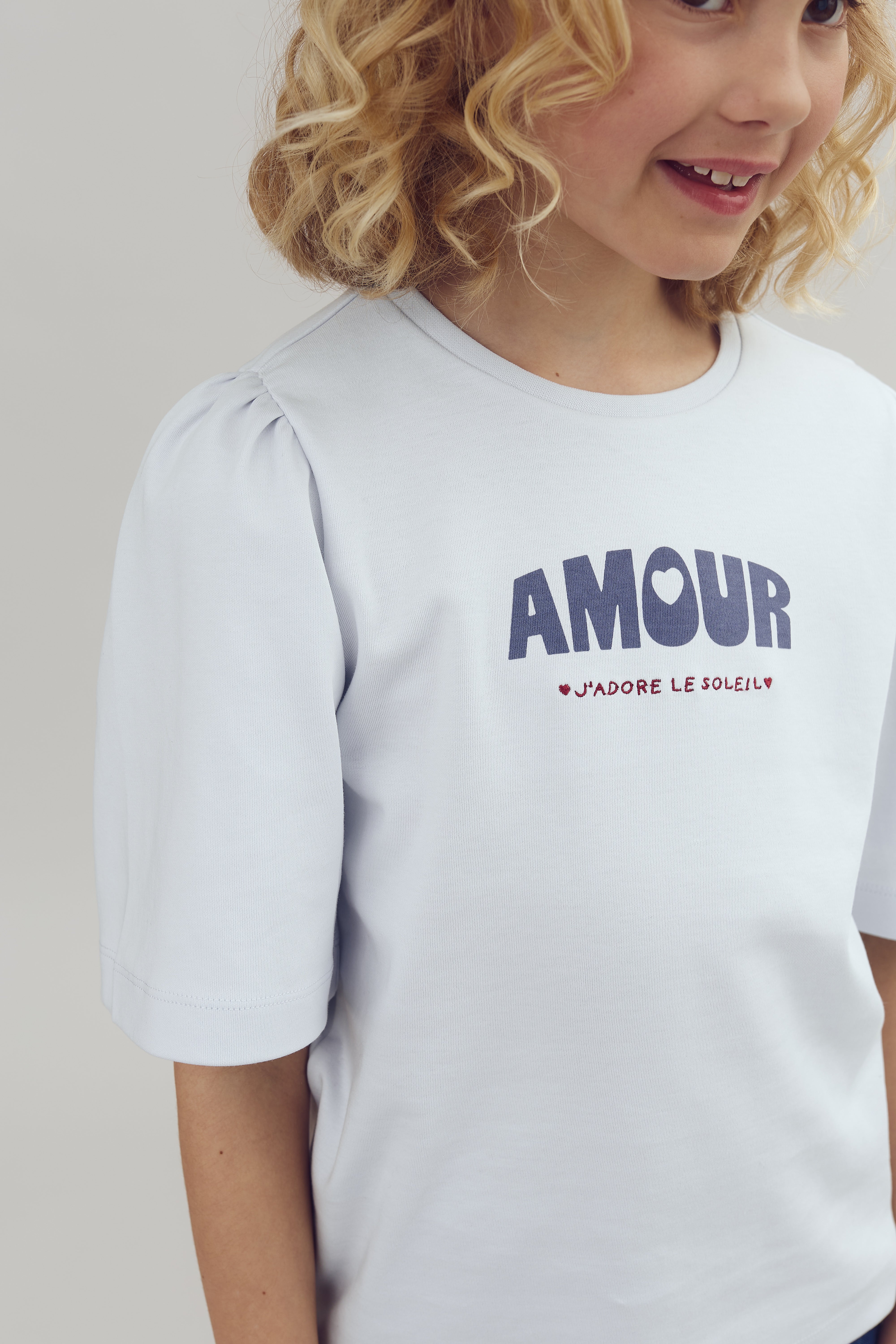Ein Kind trägt ein hellblaues T-Shirt mit den Worten "AMOUR" und "J'ADORE LE SOLEIL" darauf. Es lächelt in einer neutralen, hellen Umgebung.